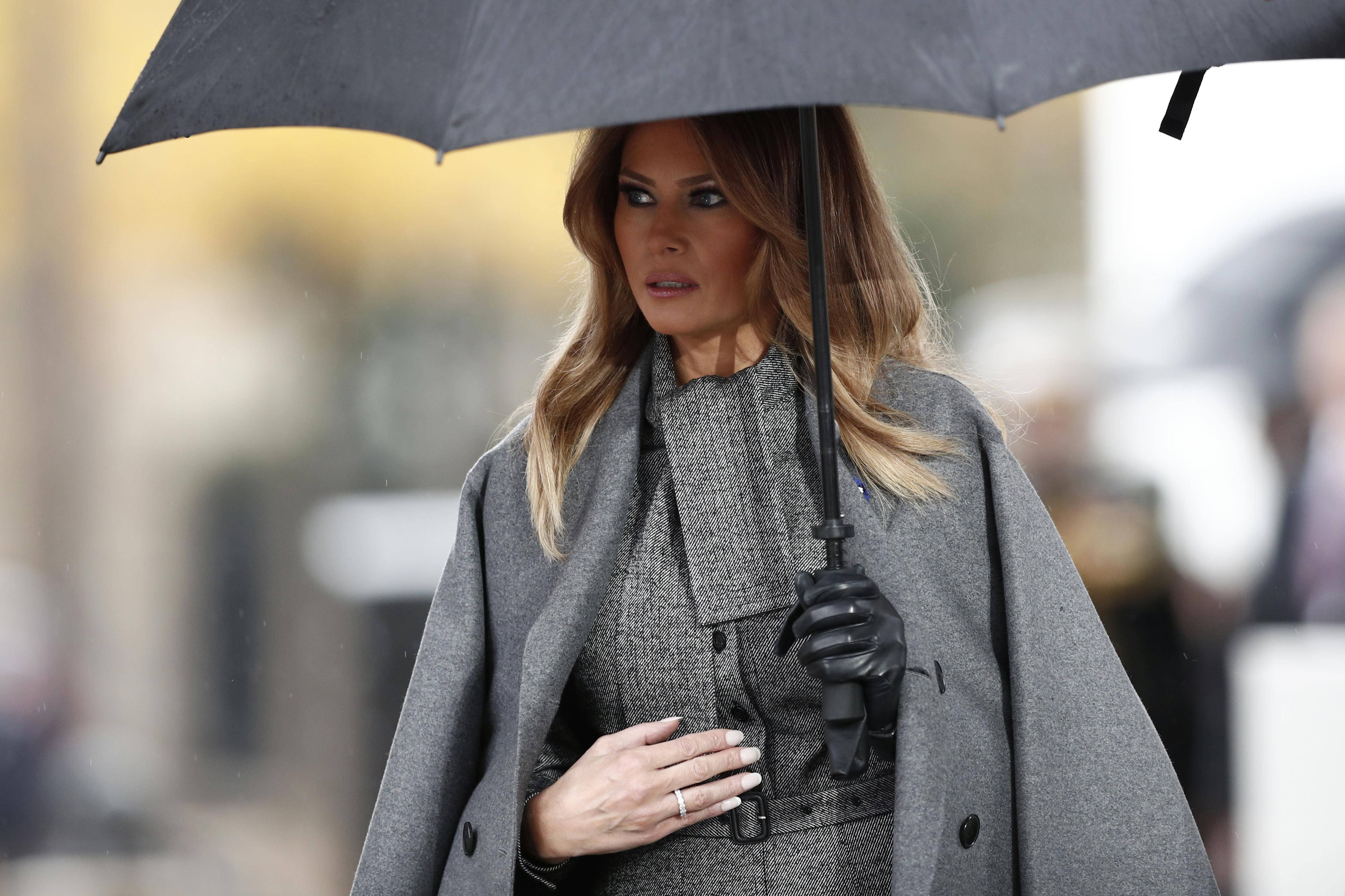Melania 桌布 壁紙 — 一位身穿灰色外套和黑色手套的女人，在雨中行走時手持一把黑色雨傘，表情嚴肅。