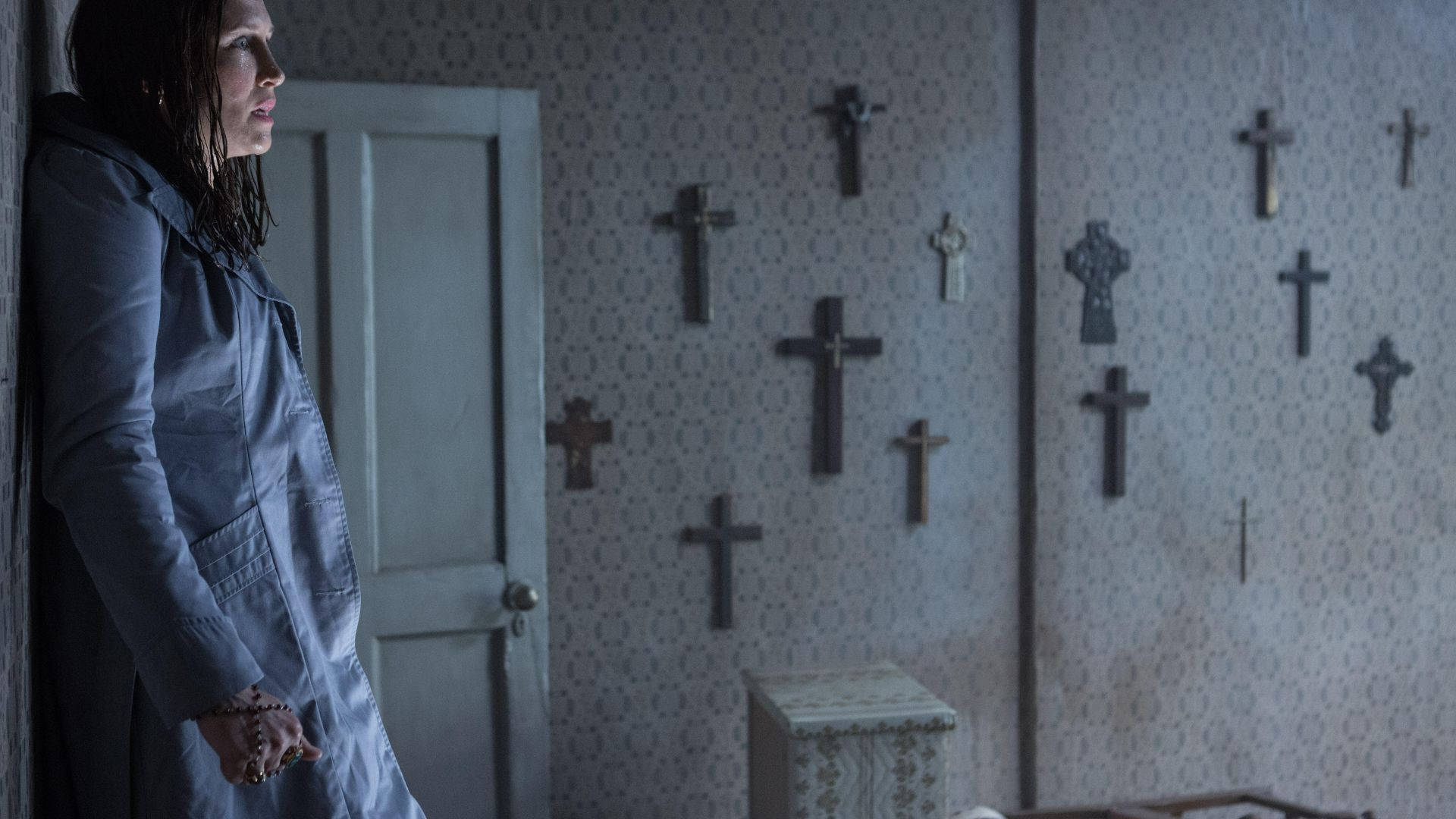 Conjuring 桌布 壁紙 — 一位身穿灰色外套的女性站在一個光線昏暗的房間裡，房間裡有圖案壁紙和許多十字架掛在牆上。