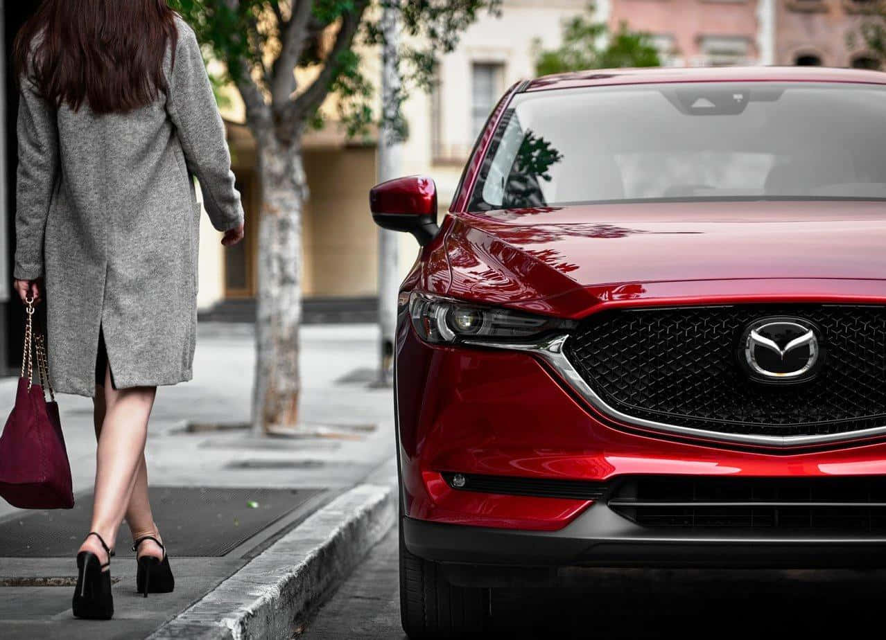Mazda Cx-5 桌布 壁紙 — 一位身穿灰色外套的女性走過一輛停在綠樹成蔭的城市街道上的閃亮紅色Mazda SUV。