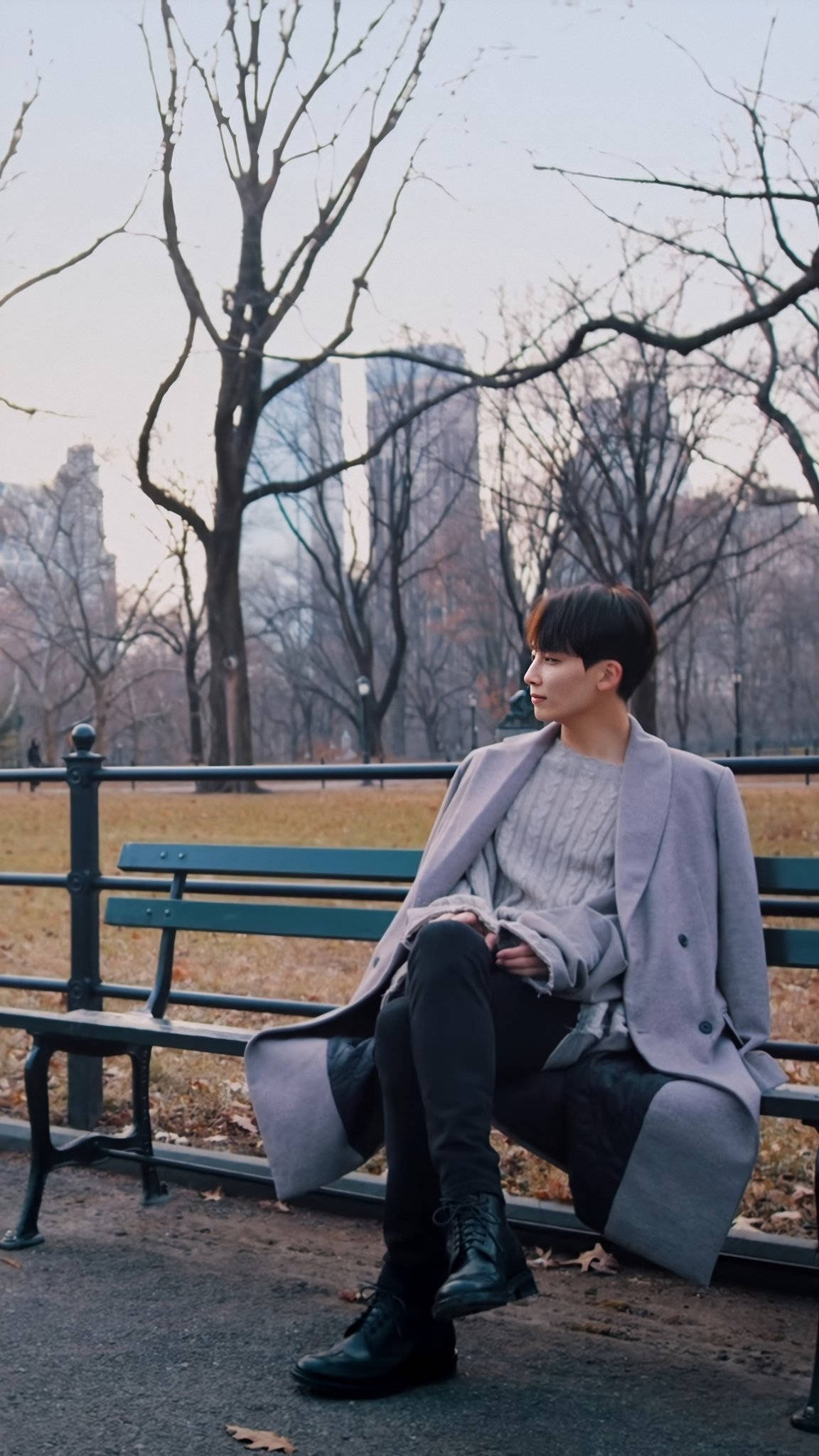 yoon jeonghan 桌布 壁紙 — 一位身穿灰色外套的年輕男子，在柔和的冬日光線中，坐在公園長椅上，周圍是光禿禿的樹木和城市摩天大樓，若有所思。