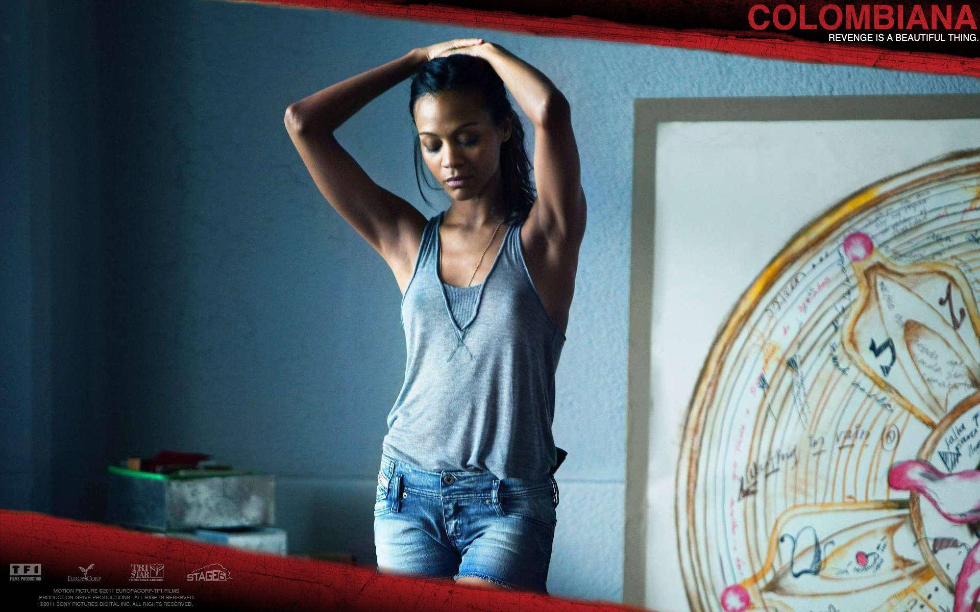 Zoe Saldana 桌布 壁紙 — 一位身穿灰色背心和牛仔短褲的女性，在光線昏暗的房間裡雙手抱頭站立，牆上掛著一個大型圓形藝術品。