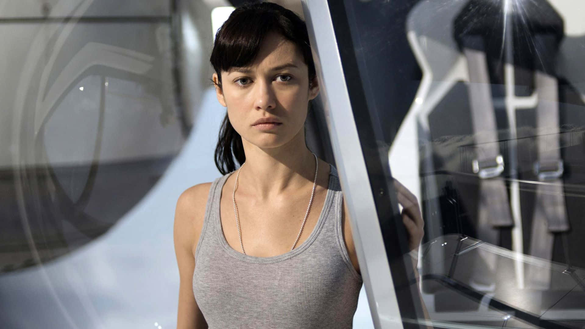 Olga Kurylenko 桌布 壁紙 — 一位身穿灰色背心的女性站在一輛具有反光表面和金屬裝飾的時尚未來感車輛旁。