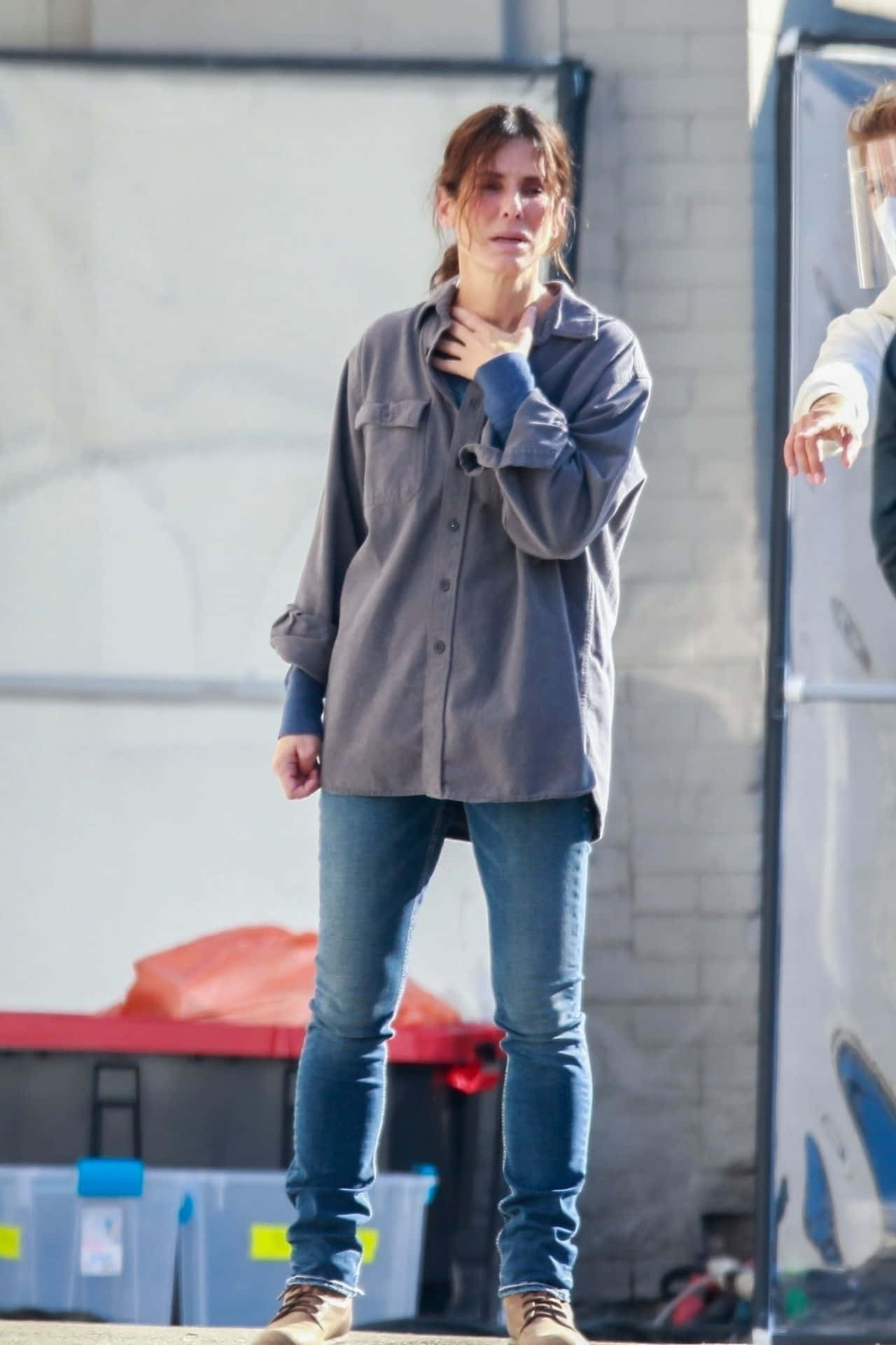 Sandra Bullock 2021 桌布 壁紙 — 一位身穿灰色襯衫和牛仔褲的女性站在戶外，表情擔憂地抓著脖子。
