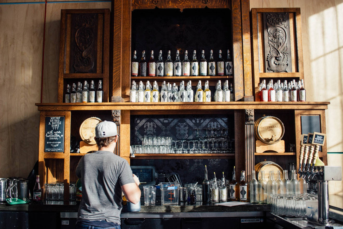 Drink 桌布 壁紙 — 一位身穿灰色襯衫和白色帽子的調酒師在擺滿酒瓶和玻璃器皿的鄉村風格木製吧檯後準備飲品。