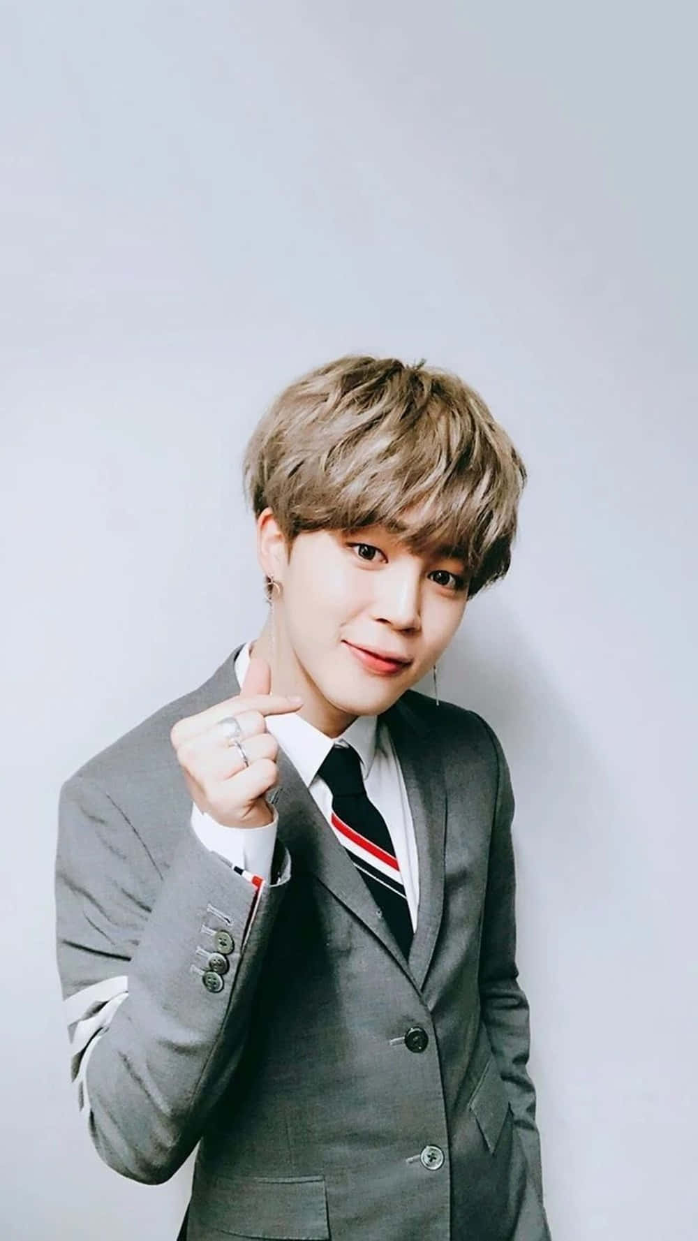 BTS 桌布 壁紙 — 一位身穿灰色西裝、打著條紋領帶的年輕男子，在純淺灰色背景前微笑著指向觀看者。