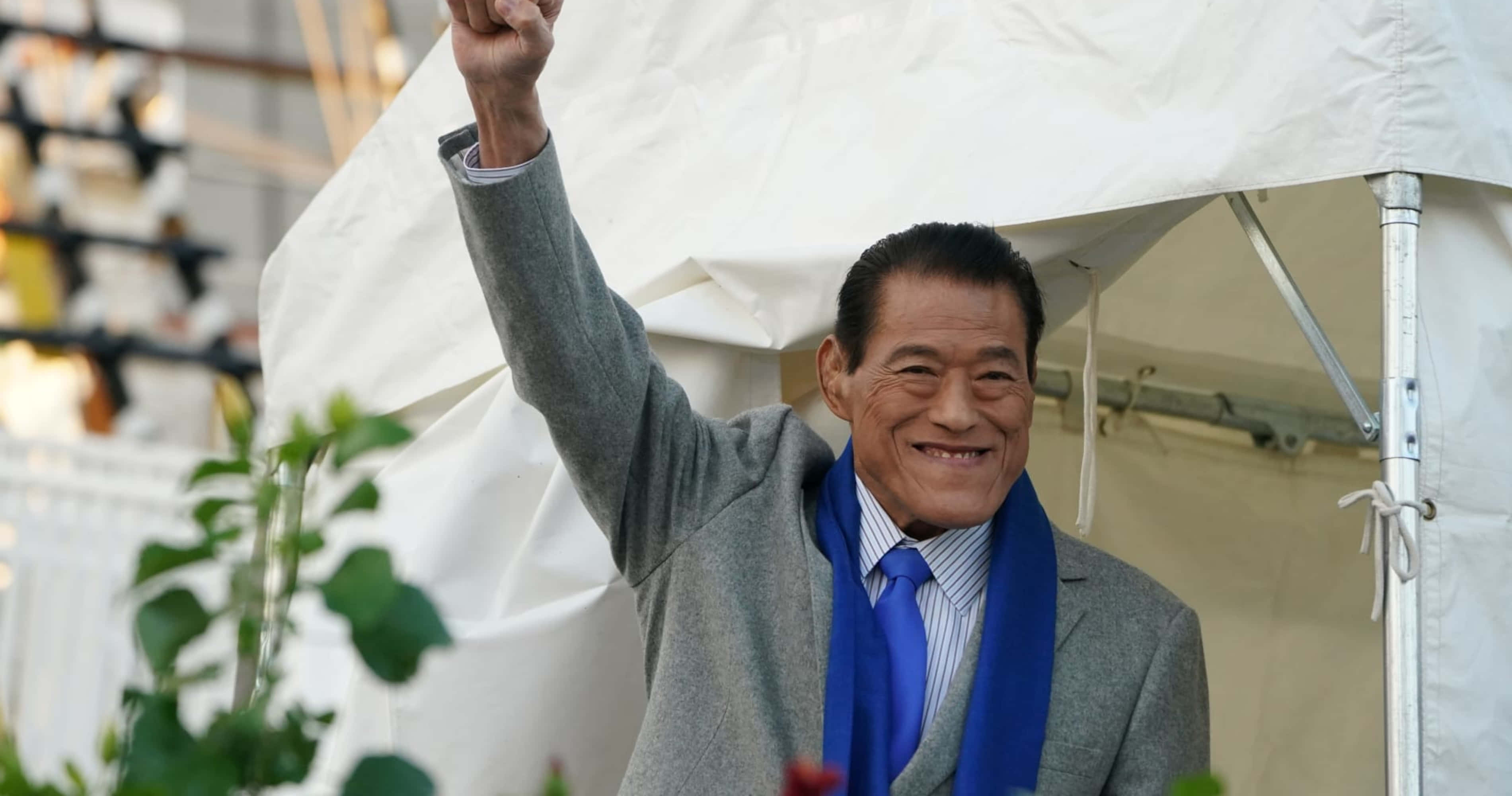 摔角 桌布 壁紙 — 一位身穿灰色西裝和藍色領帶的年長男性在白色帳篷下微笑著舉起拳頭。