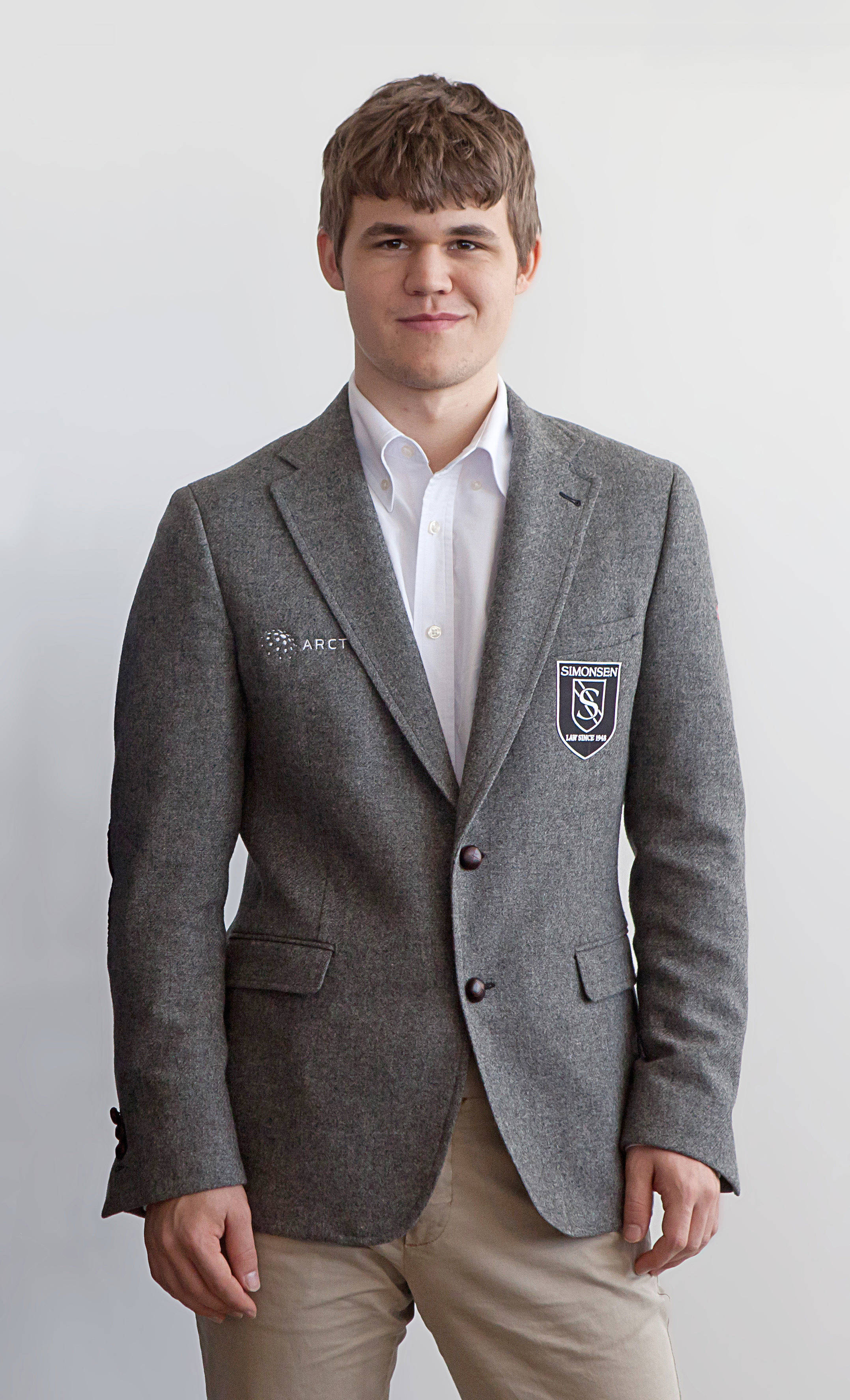 Magnus 桌布 壁紙 — 一位身穿灰色西裝外套（帶有刺繡補丁）的年輕男子，自信地站在純白色背景前。