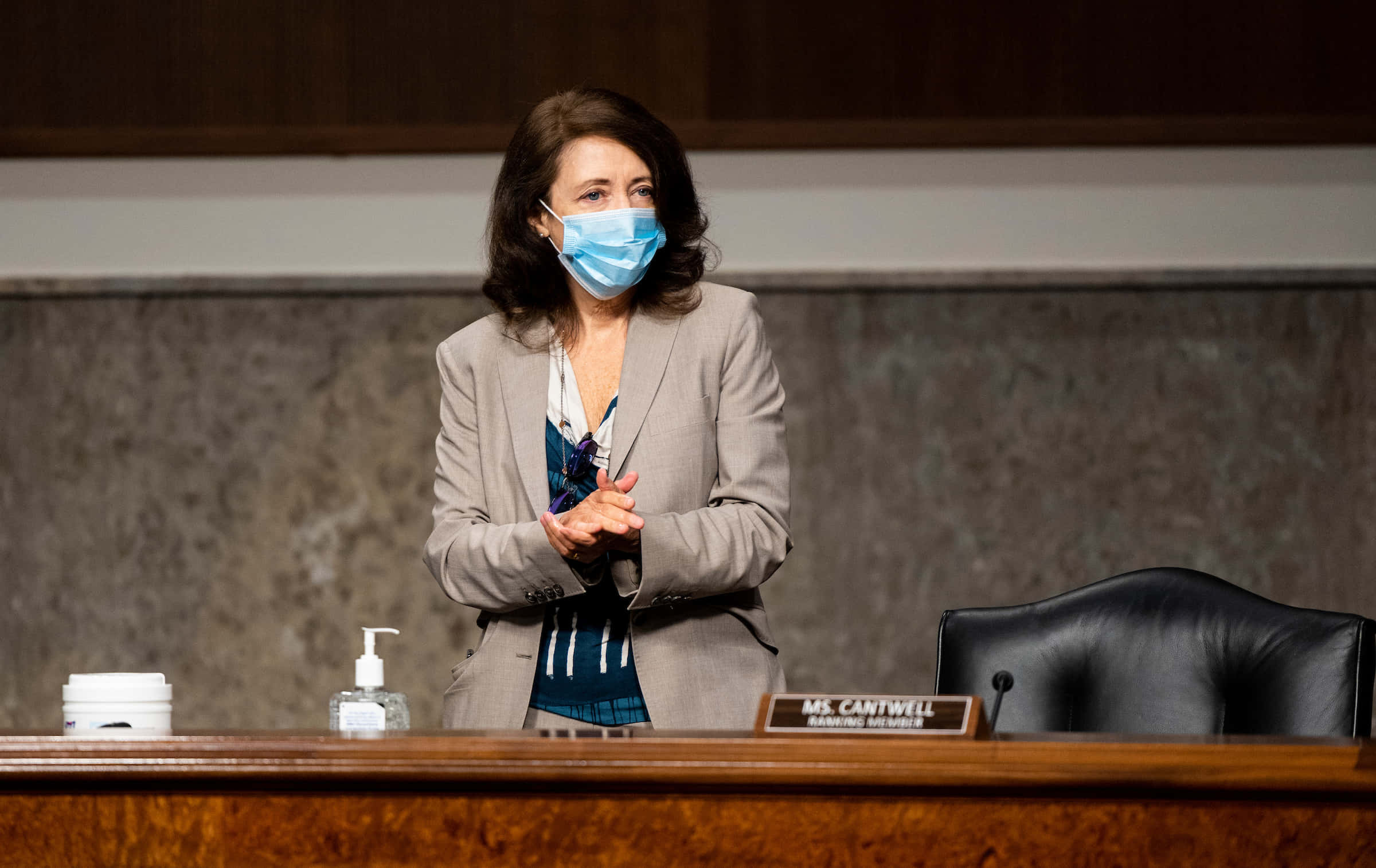政治 桌布 壁紙 — 一位身穿灰色西裝外套和藍色口罩的女性站在一張木桌前，桌上有一個寫著Ms Cantwell的名牌，位於正式的聽證室內。