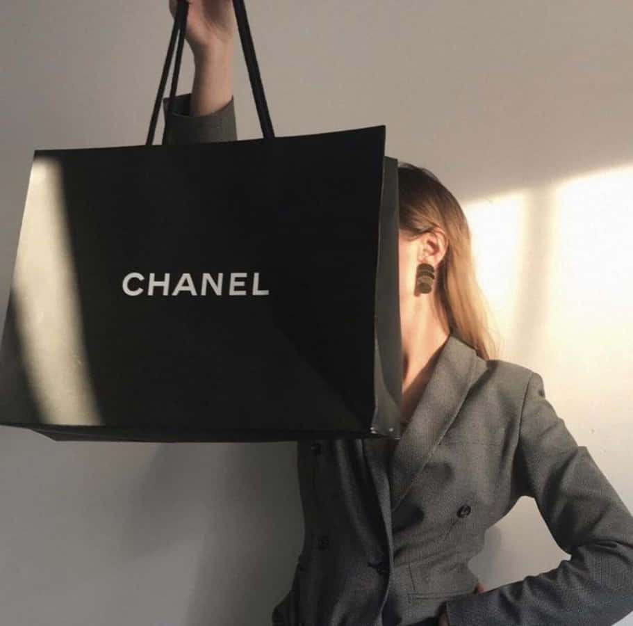 Chanel 桌布 壁紙 — 一位身穿灰色西裝外套的女性將一個大型黑色Chanel購物袋舉過臉部，部分遮蓋了她的容貌，陽光在她身後的一面素牆上投下陰影。