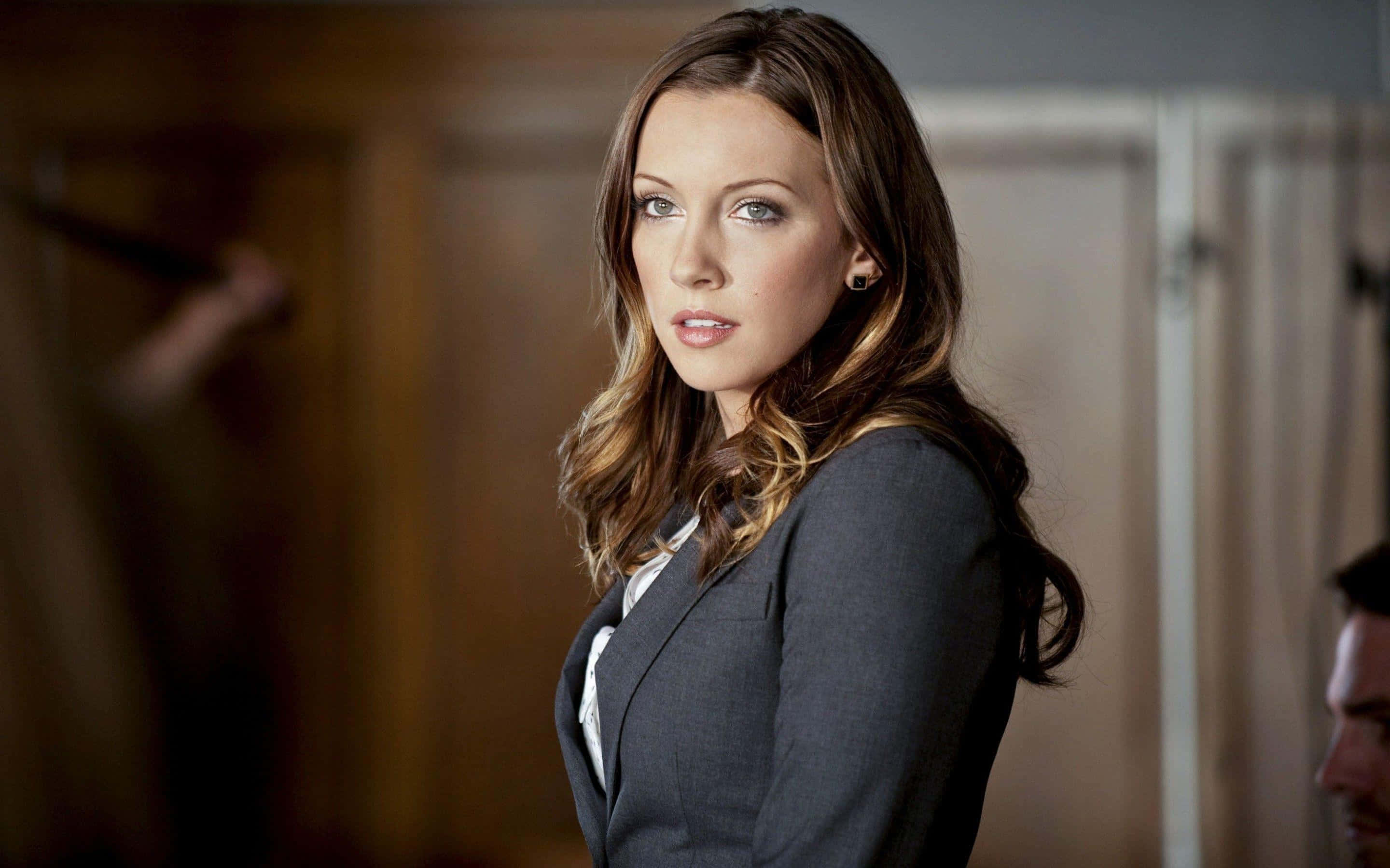 katie cassidy 桌布 壁紙 — 一位身穿灰色西裝外套的女性站在燈光昏暗的房間裡，表情嚴肅，柔和的光線凸顯了她的五官。