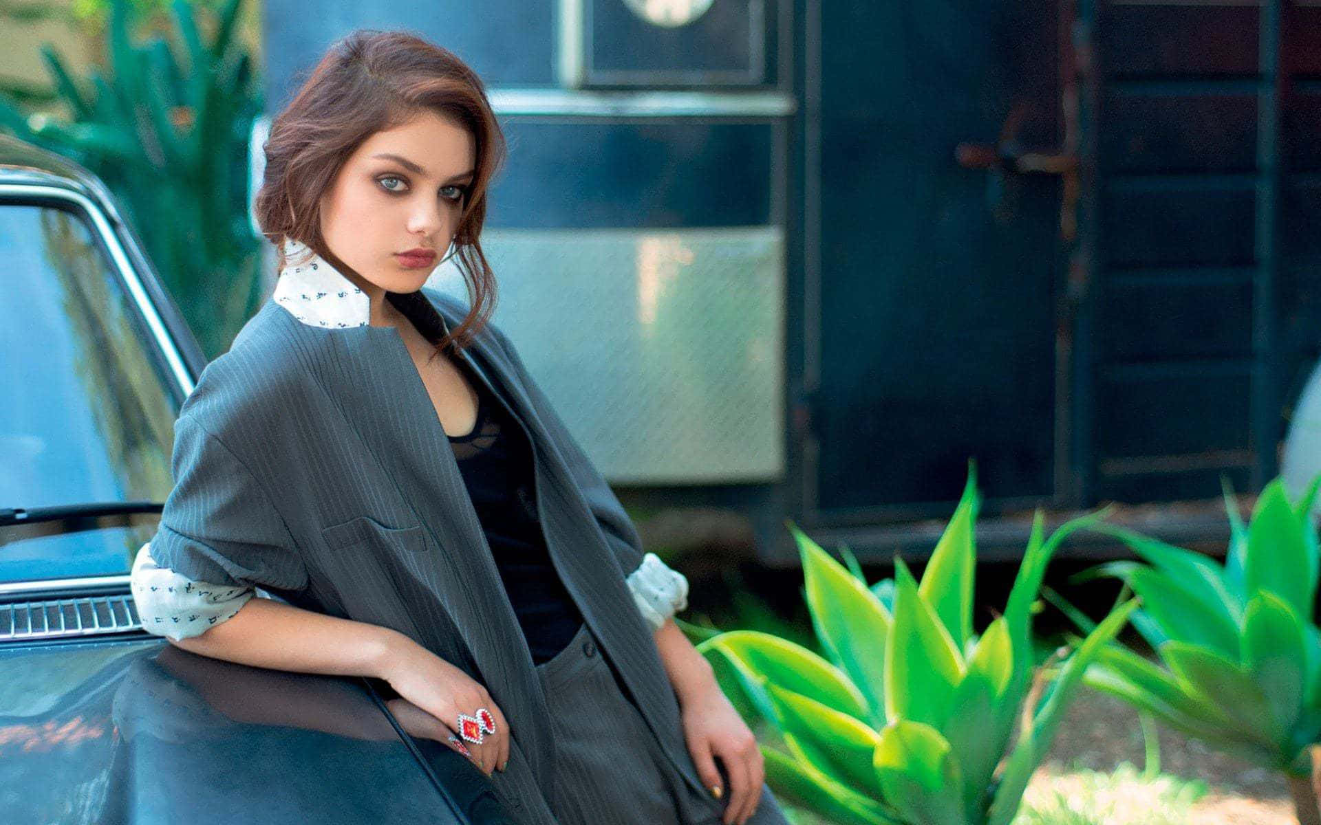 Odeya Rush 桌布 壁紙 — 一位身穿灰色西裝外套的女性靠在一輛復古車上，前景有充滿活力的綠色多肉植物。