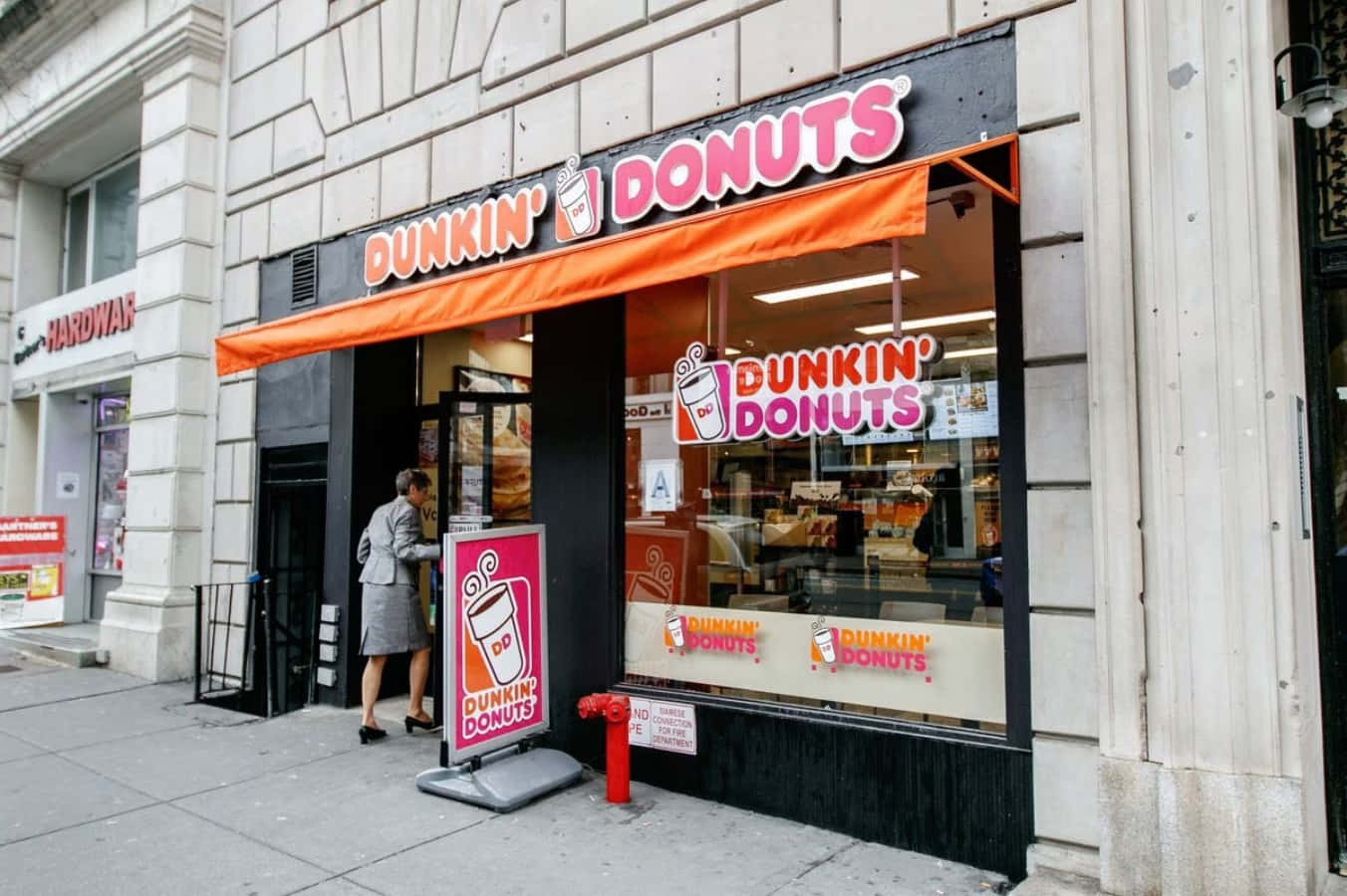 Dunkin Donuts 桌布 壁紙 — 一位身穿灰色西裝的女士在城市人行道上走過一間Dunkin' Donuts店鋪前鮮橙色的遮陽篷。