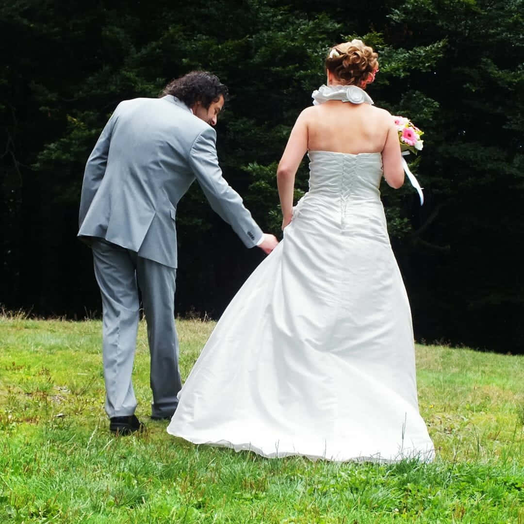 Wedding 桌布 壁紙 — 一位身穿灰色西裝的新郎輕輕掀起新娘白色婚紗的裙擺，他們站在草地上，身後有深色的樹木。