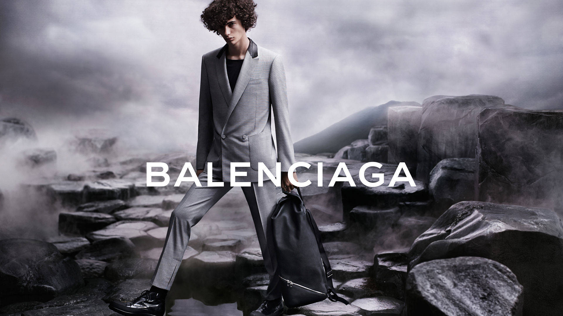 Balenciaga 桌布 壁紙 — 一位身穿灰色西裝的模特兒在風暴天空下自信地走過霧氣瀰漫的岩石地形，並疊加了「BALENCIAGA」字樣。