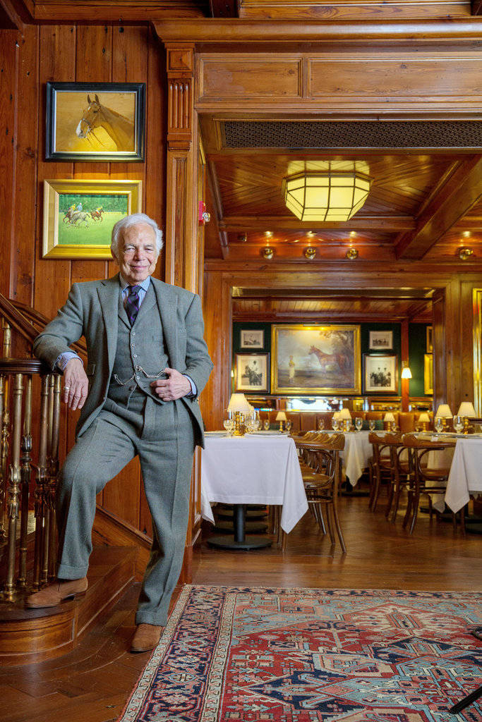 ralph lauren 桌布 壁紙 — 一位身穿灰色西裝的長者，在燈光溫暖、以木鑲板裝飾的餐廳中，隨意地靠在木樓梯上，地上鋪有圖案地毯，牆上掛著裱框的馬術藝術品。