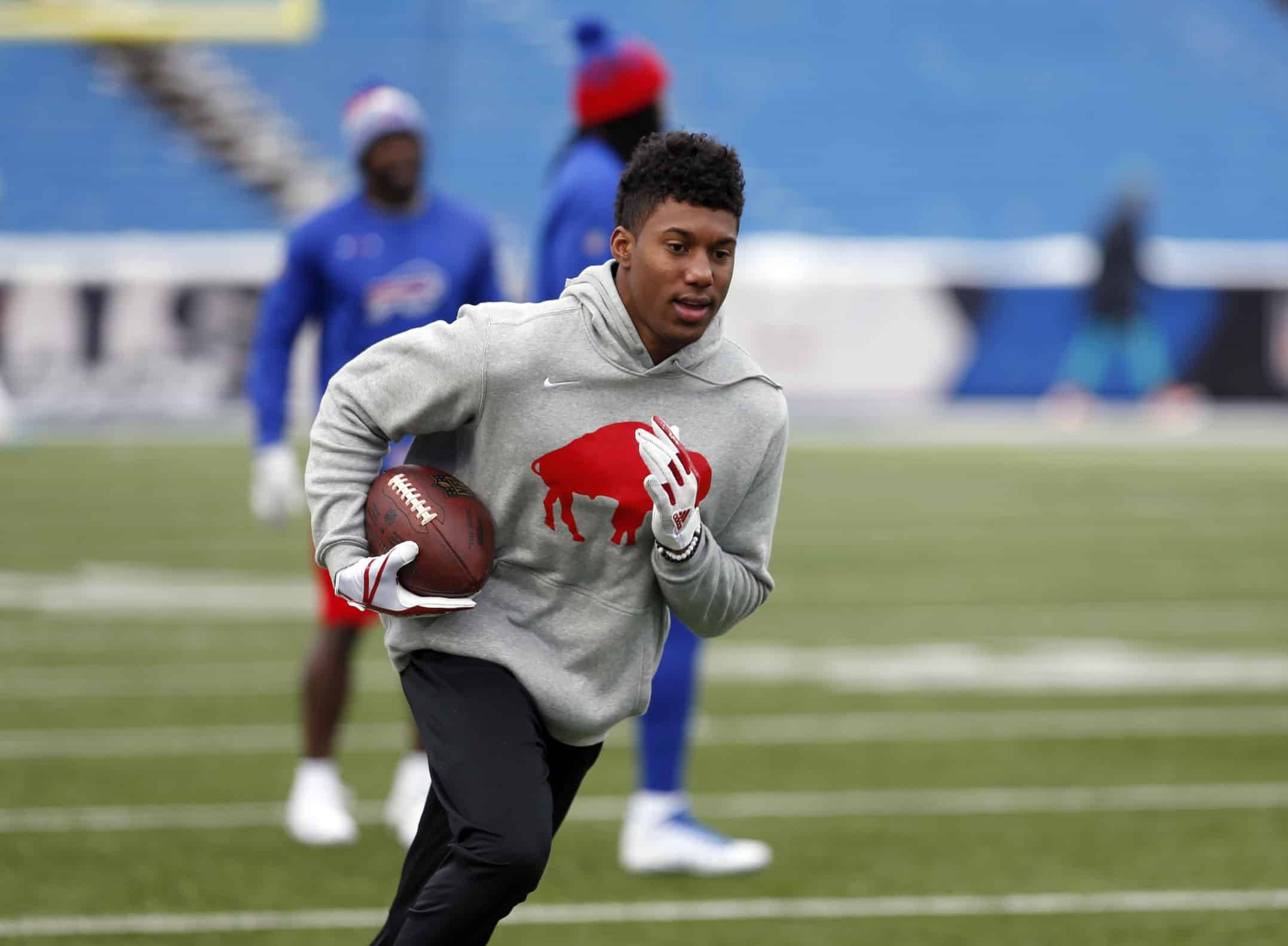 zay jones 桌布 壁紙 — 一位身穿灰色連帽衫、帶有紅色水牛標誌的年輕美式足球員，手持棕色美式足球在綠色球場上衝刺。