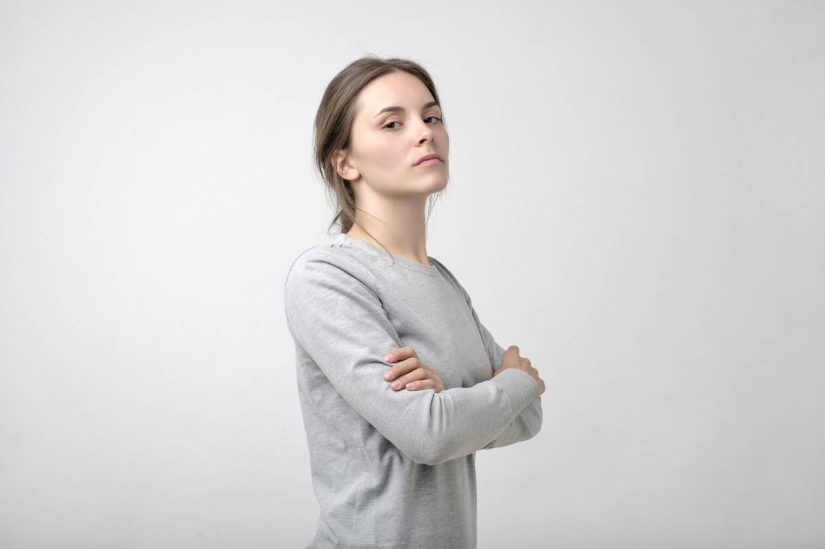 模特兒 桌布 壁紙 — 一位身穿灰色運動衫的女性，雙臂交叉，站在純白色背景前，表情嚴肅地看向一旁。