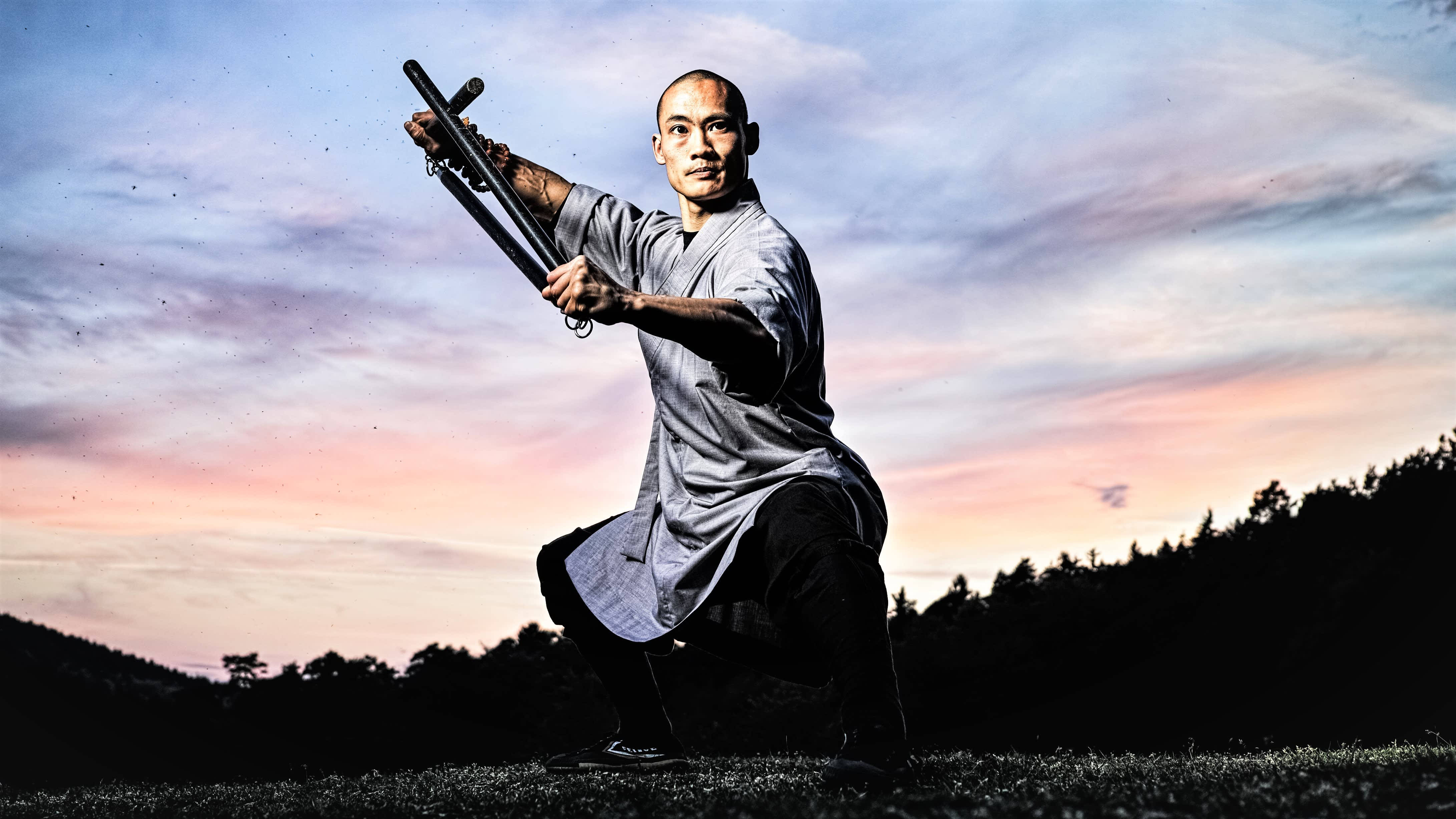 martial arts 桌布 壁紙 — 一位身穿灰色長袍的武術家，在充滿活力的暮色天空下，用雙節棍擺出強而有力的姿勢。