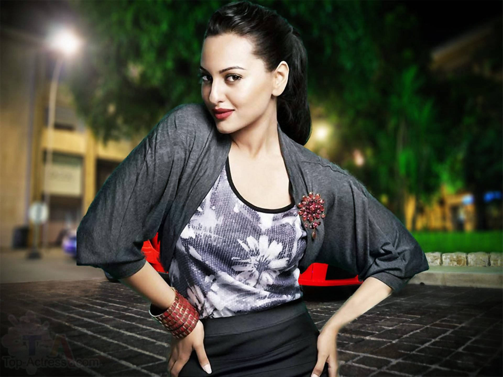 Sonakshi Sinha 桌布 壁紙 — 一位身穿灰色開襟衫和花卉上衣的女性，在夜晚的路燈下自信地站在磚砌人行道上擺姿勢。