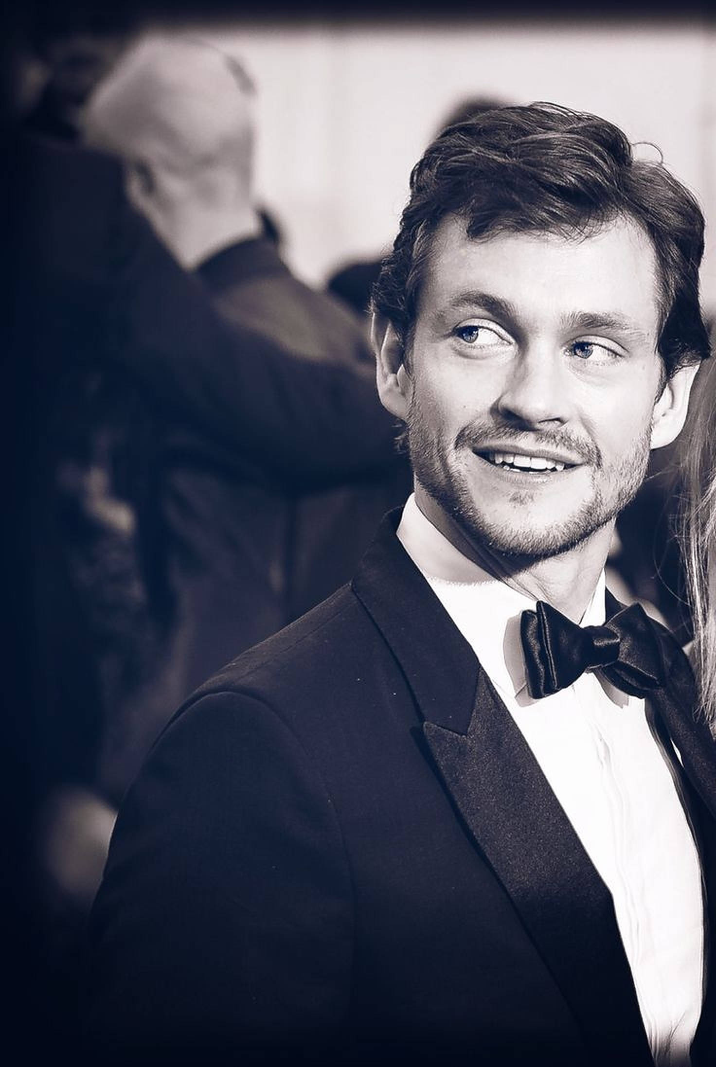 Hugh Dancy 桌布 壁紙 — 一位身穿燕尾服、打著蝴蝶結的微笑男士在淺景深黑白肖像中望向鏡頭外。