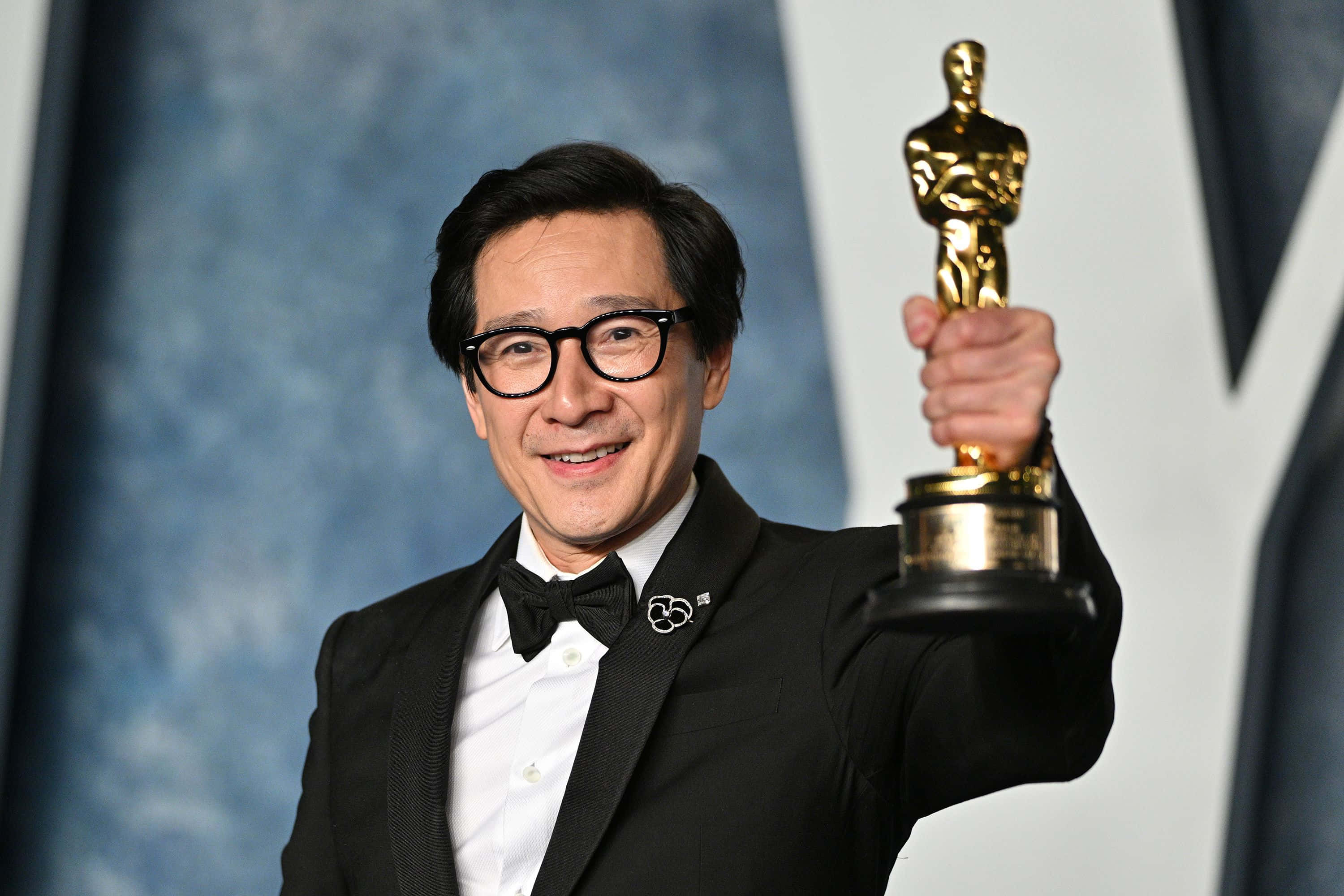 Ke Huy Quan 桌布 壁紙 — 一位身穿燕尾服、面帶微笑的男子，在藍色紋理背景前自豪地舉起一座金色奧斯卡獎座。