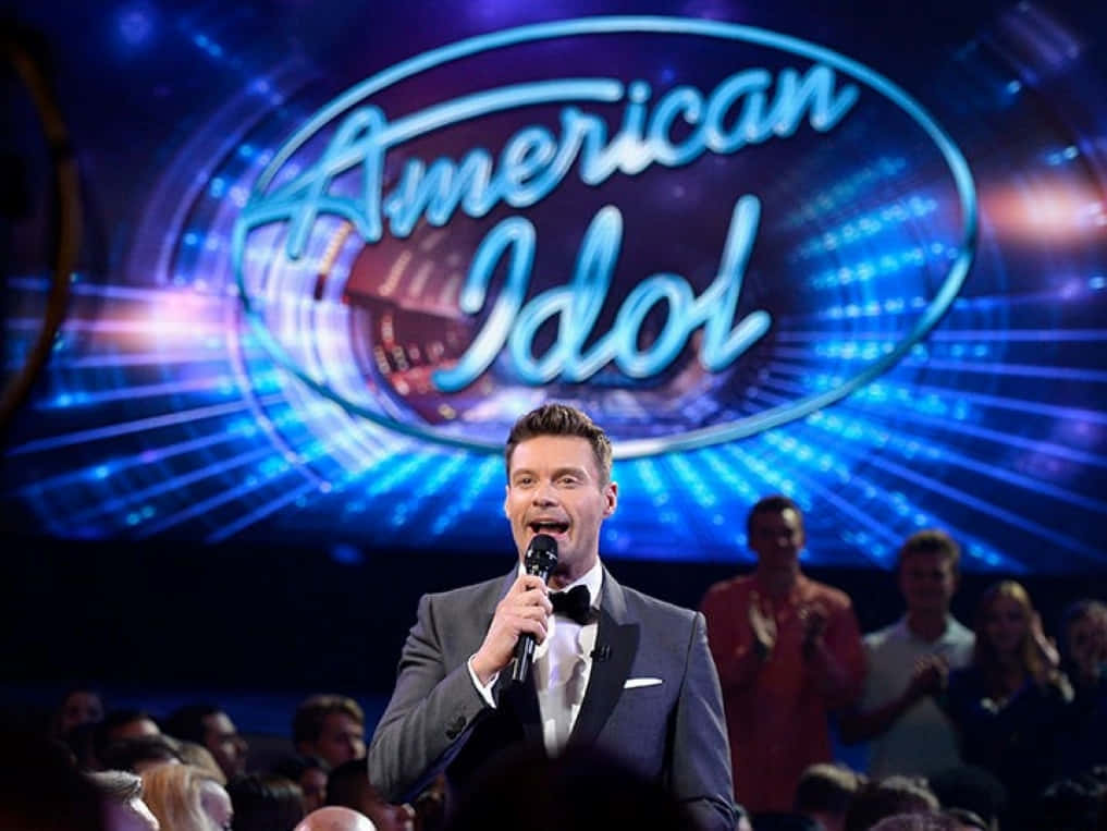 american idol 桌布 壁紙 — 一位身穿燕尾服的主持人在燈光明亮的美國偶像舞台上，對著麥克風唱歌，背景有發光的藍色燈光和觀眾。