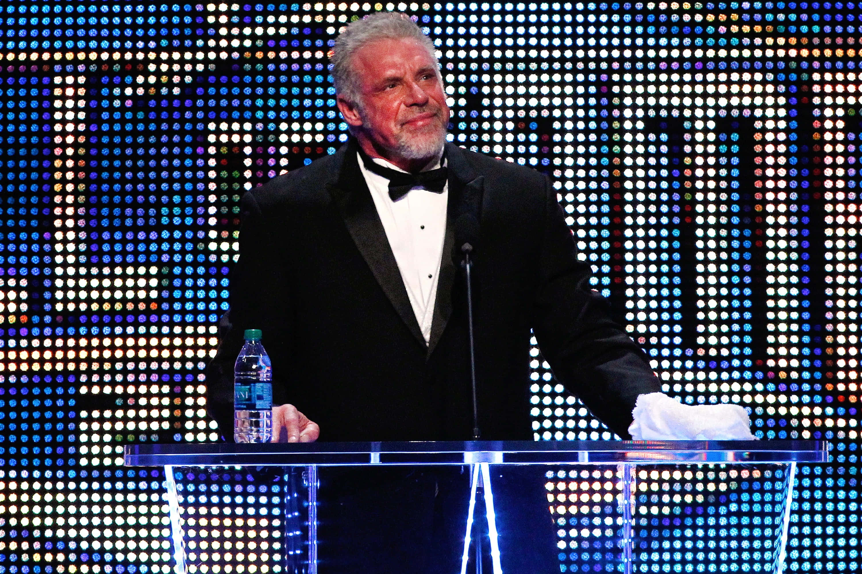 WWE 桌布 壁紙 — 一位身穿燕尾服的男士站在透明講台前，背景是耀眼的多彩燈光網格。