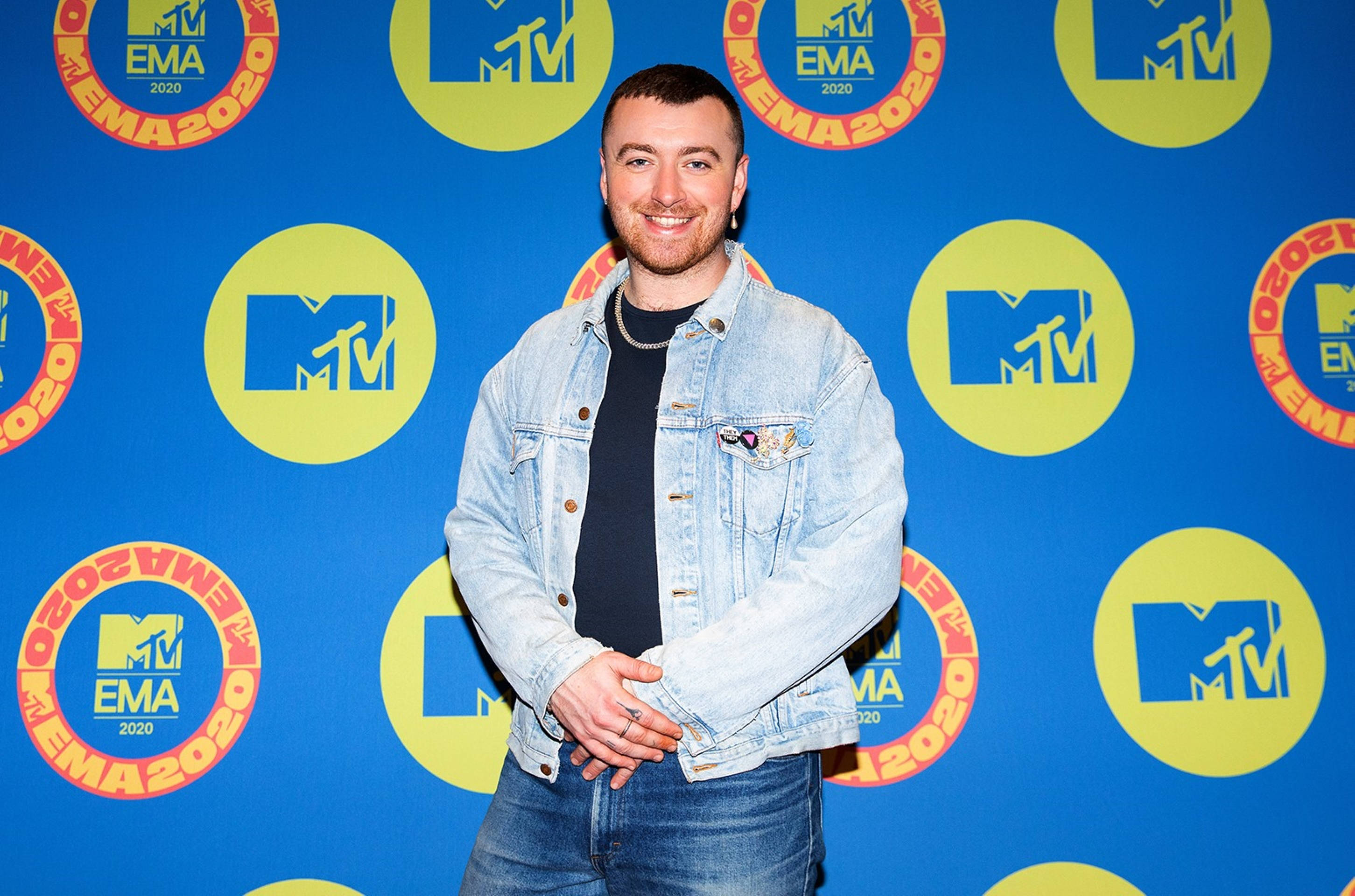 Sam Smith 桌布 壁紙 — 一位身穿牛仔外套、面帶微笑的男子自信地站在充滿活力的藍色背景前，背景上重複出現MTV EMA 2020標誌。