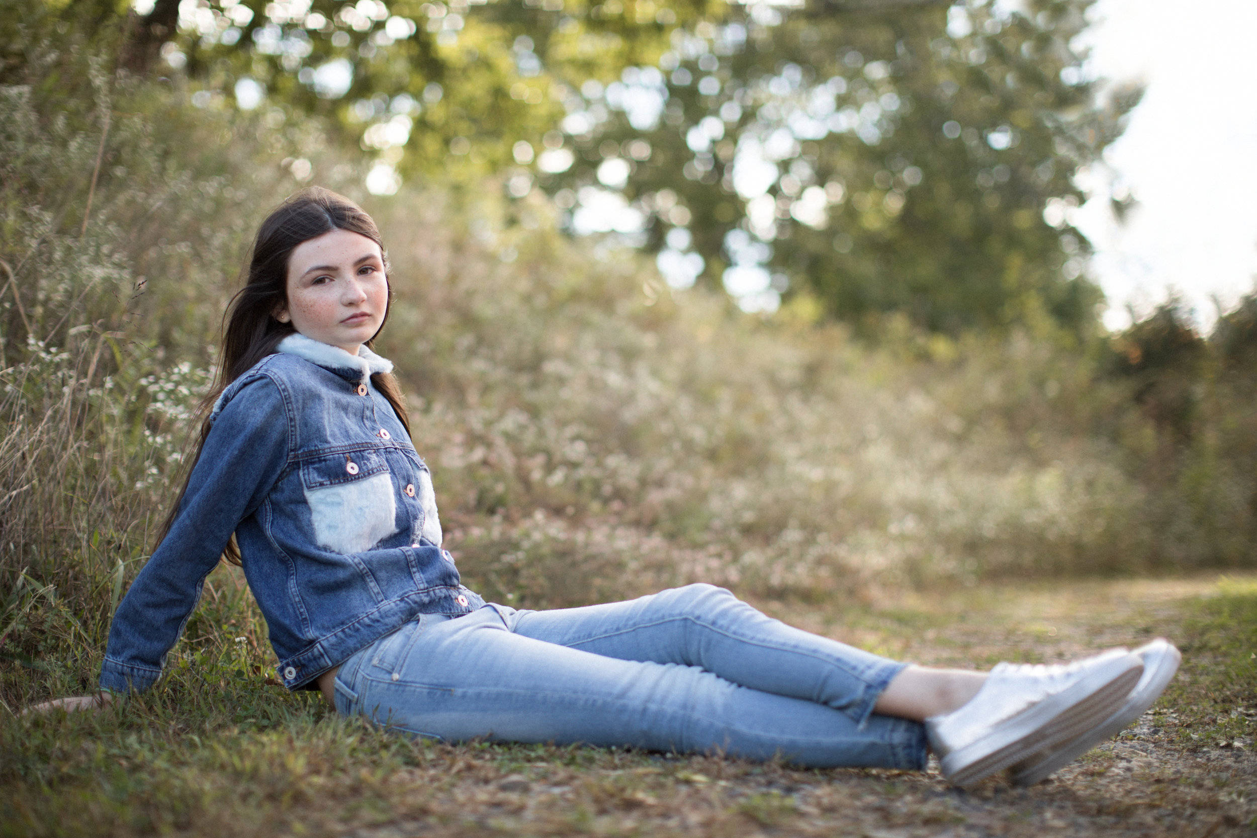 Denim 桌布 壁紙 — 一位身穿牛仔外套和牛仔褲的年輕女性，在柔和背景的樹木下，斑駁的陽光中放鬆地坐在草地上。