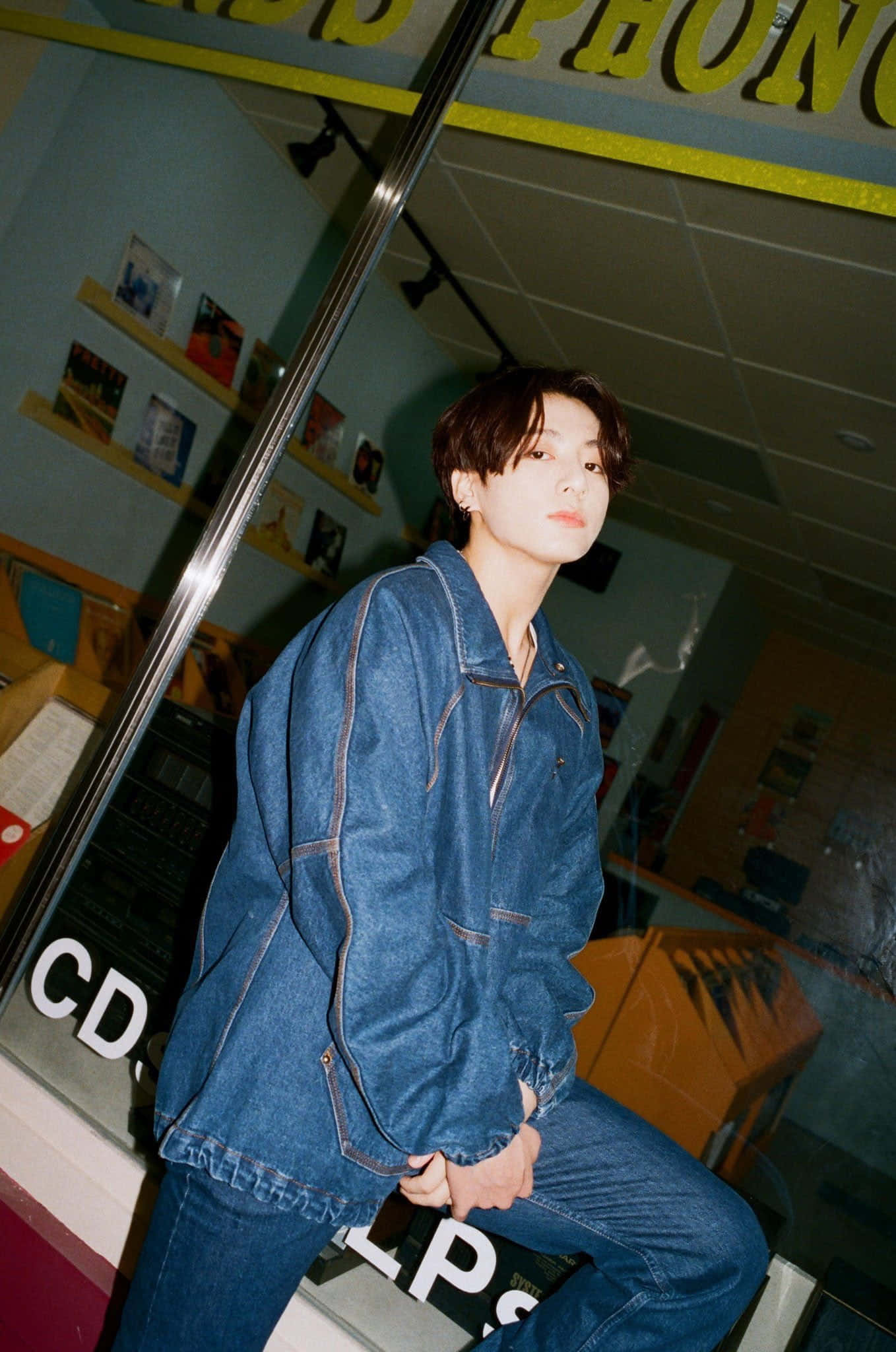 Jungkook 桌布 壁紙 — 一位身穿牛仔外套和牛仔褲的年輕男子靠在一間復古唱片行的玻璃展示櫃上，店內有色彩繽紛的貨架和霓虹燈招牌。