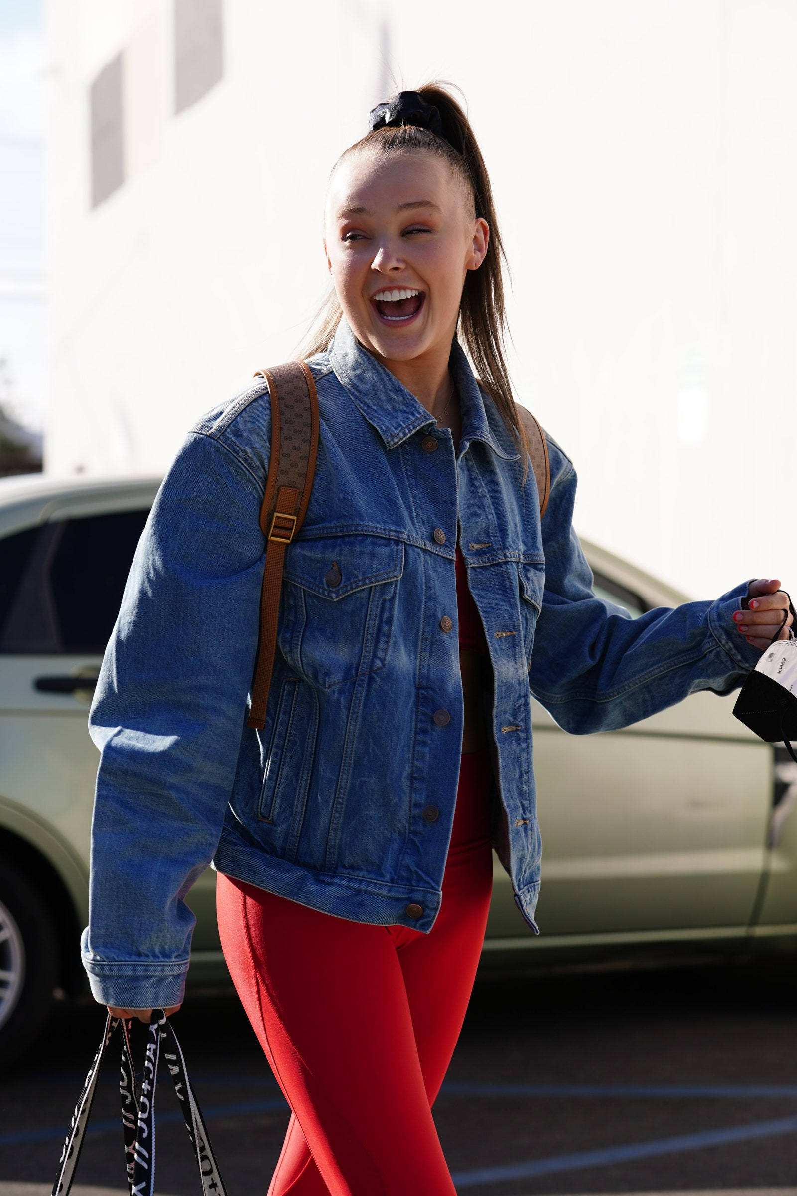 JoJo Siwa 桌布 壁紙 — 一位身穿牛仔外套和紅色褲子的年輕女子，帶著購物袋在戶外快樂地笑著走路。