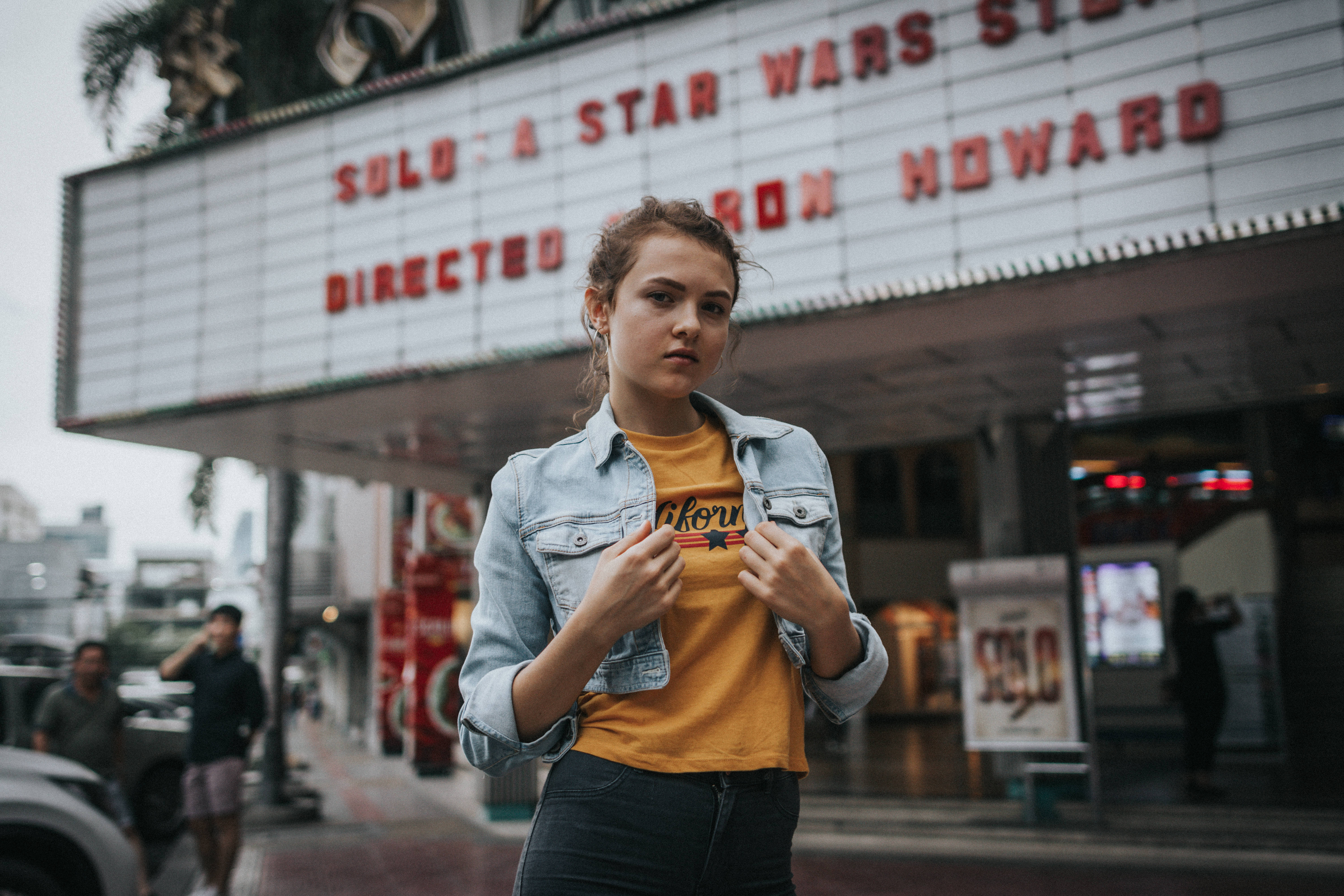 vintage star wars iPhone 桌布 壁紙 — 一位身穿牛仔外套和黃色T恤的年輕女性自信地站在電影院簷篷前，上面顯示著「Solo A Star Wars Story」。
