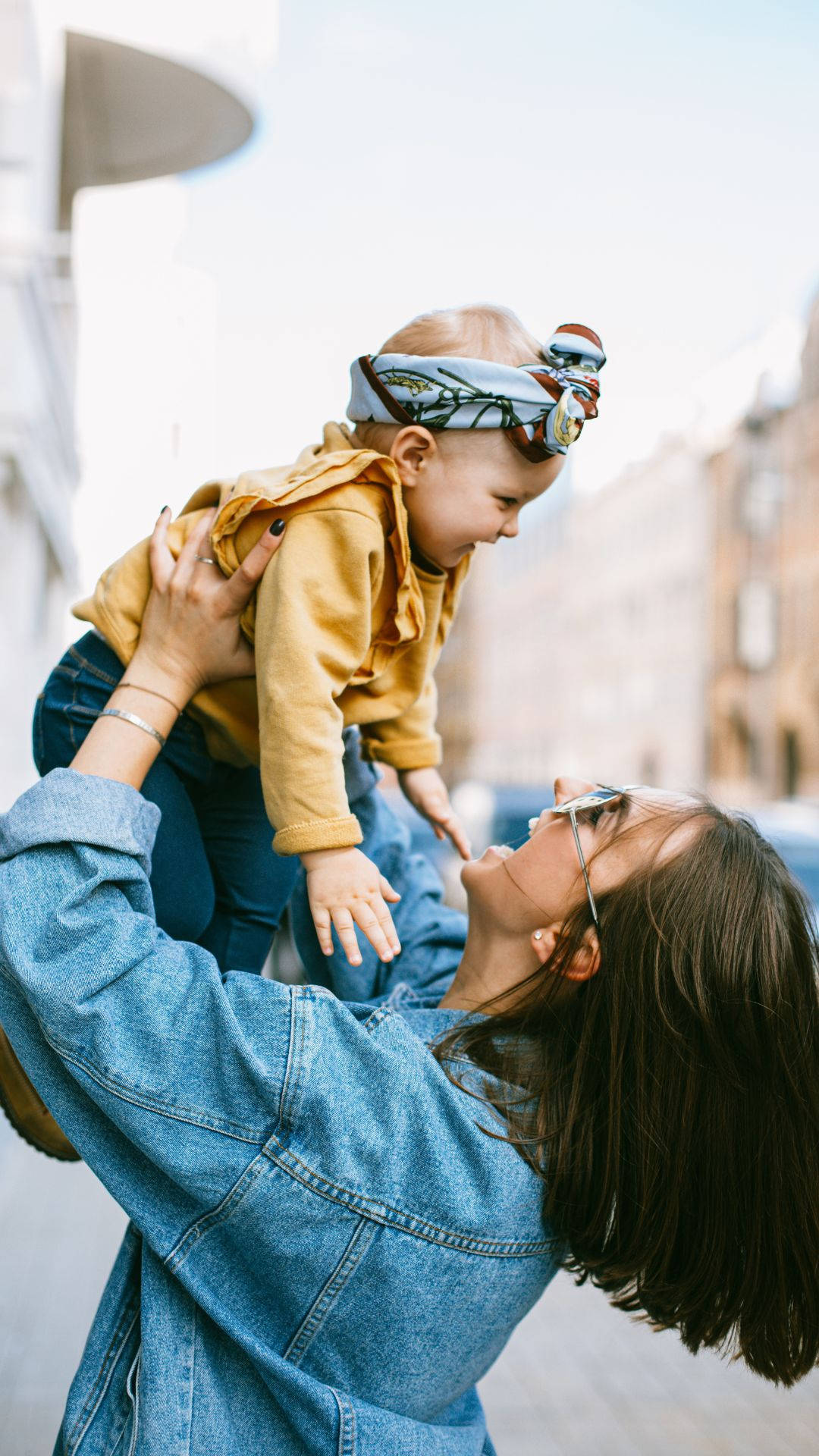 Baby 桌布 壁紙 — 一位身穿牛仔外套的女性在戶外抱起一個戴著圖案頭帶和黃色連帽衫的嬰兒。
