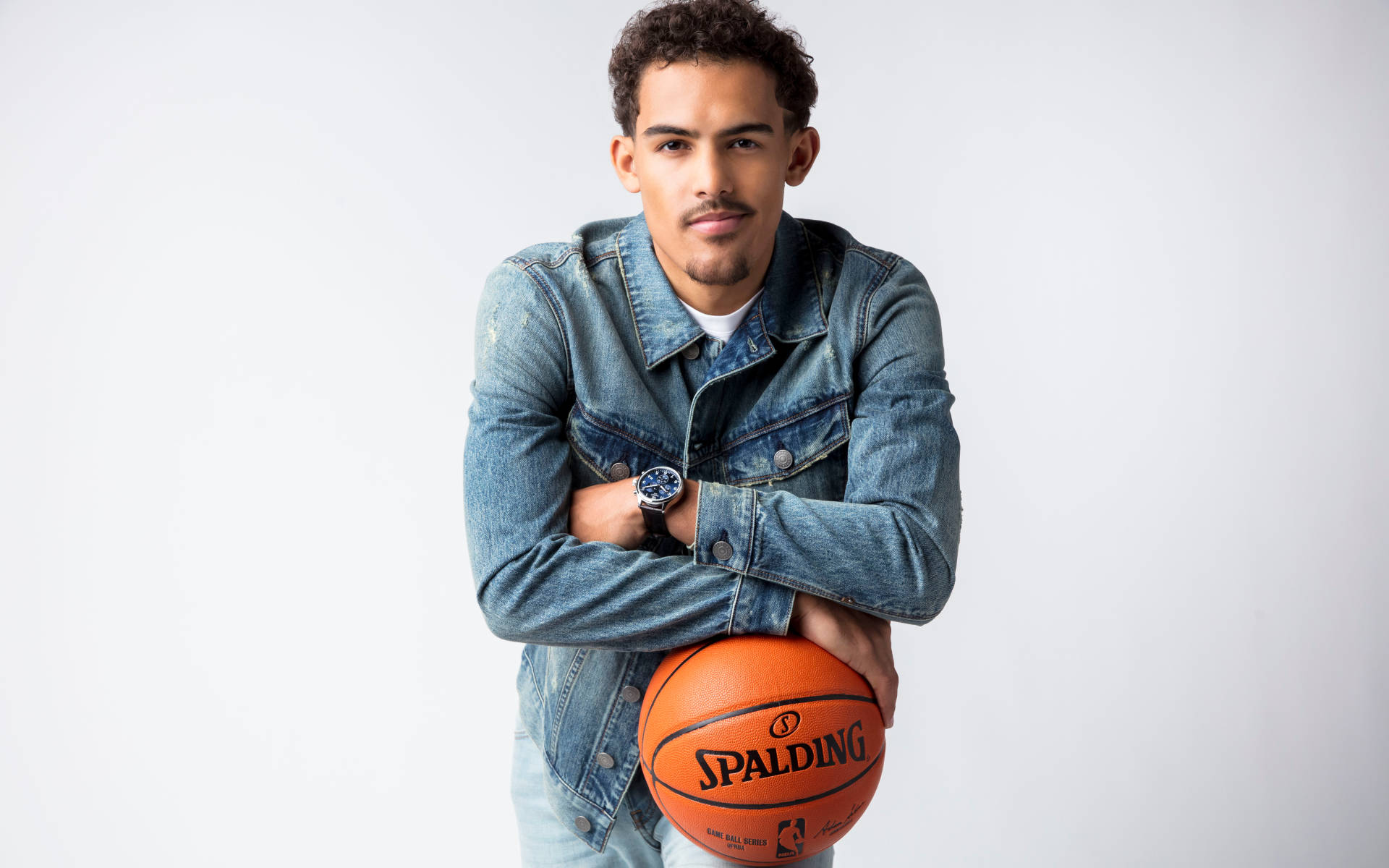 Trae Young 桌布 壁紙 — 一位身穿牛仔外套的年輕男子，在純白色背景前拿著一個橙色的 Spalding 籃球。