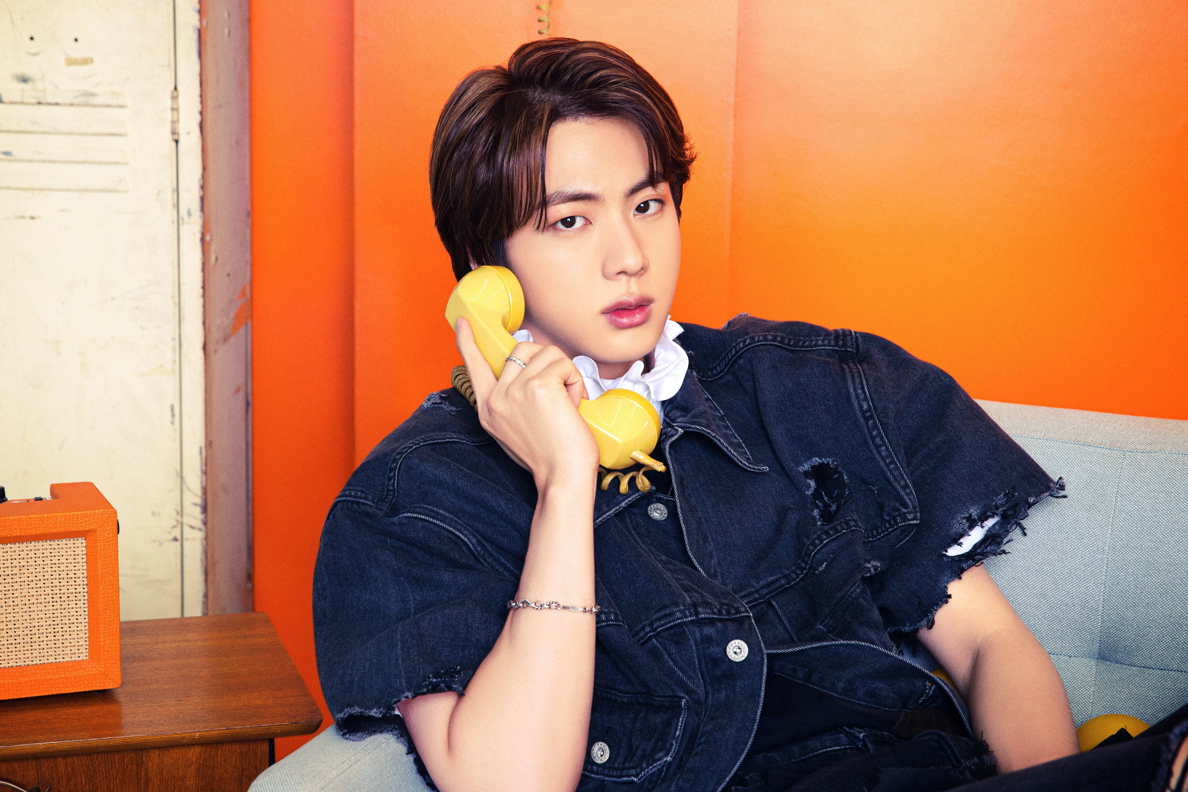 kim seok jin 桌布 壁紙 — 一位身穿牛仔外套的年輕男子，坐在沙發上，對著鮮橙色牆壁，將黃色轉盤電話貼在耳邊。