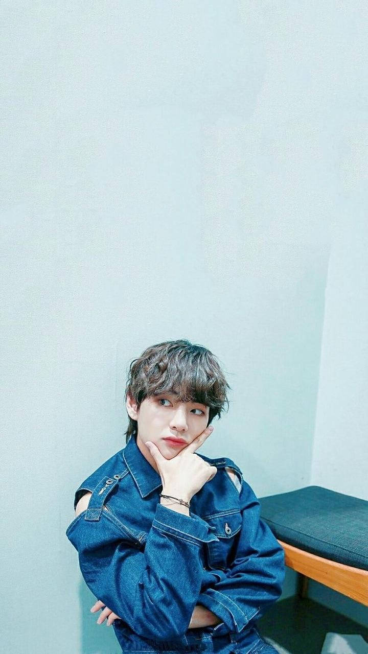 Taehyung Cute 桌布 壁紙 — 一位身穿牛仔外套的年輕男子，將下巴擱在手上，靠著一面淺藍色素牆。