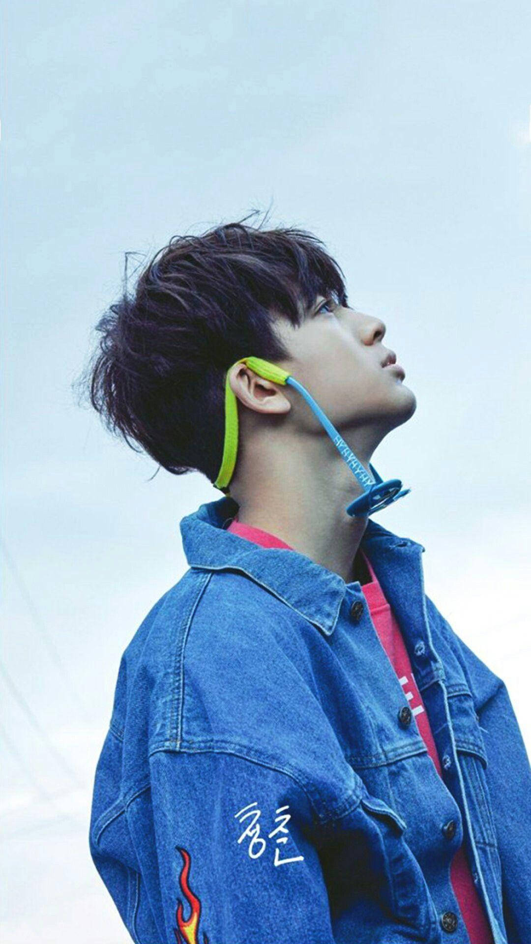 Ikon 桌布 壁紙 — 一位身穿牛仔外套的年輕男子，脖子上掛著彩色耳機，仰望蒼白的天空。