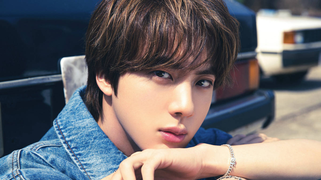 kim seok jin 桌布 壁紙 — 一位身穿牛仔外套的年輕男子在明亮陽光下，將下巴擱在手上，旁邊有一輛經典汽車。