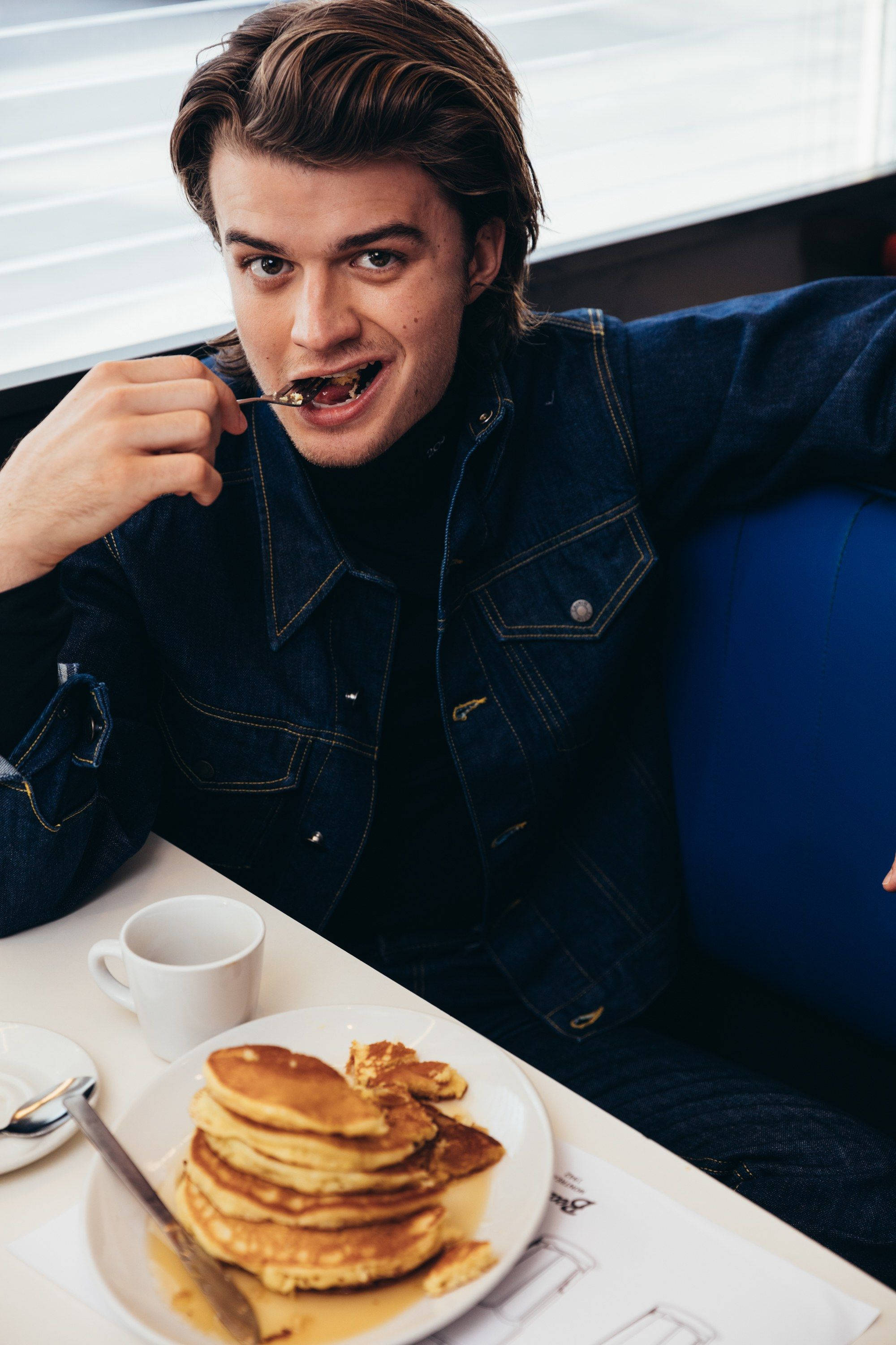 Joe Keery 桌布 壁紙 — 一位身穿牛仔外套的年輕男子在餐廳卡座吃鬆餅，身後是明亮的窗戶。