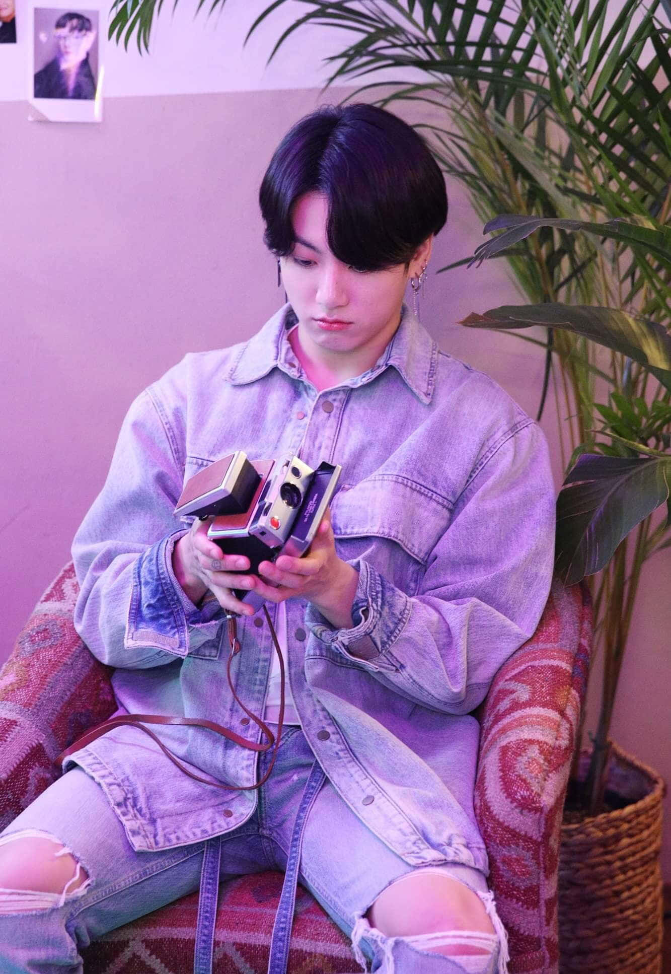 Jungkook 桌布 壁紙 — 一位身穿牛仔外套的年輕男子坐在圖案椅子上，在紫色燈光下拿著復古相機，旁邊有一盆盆栽。
