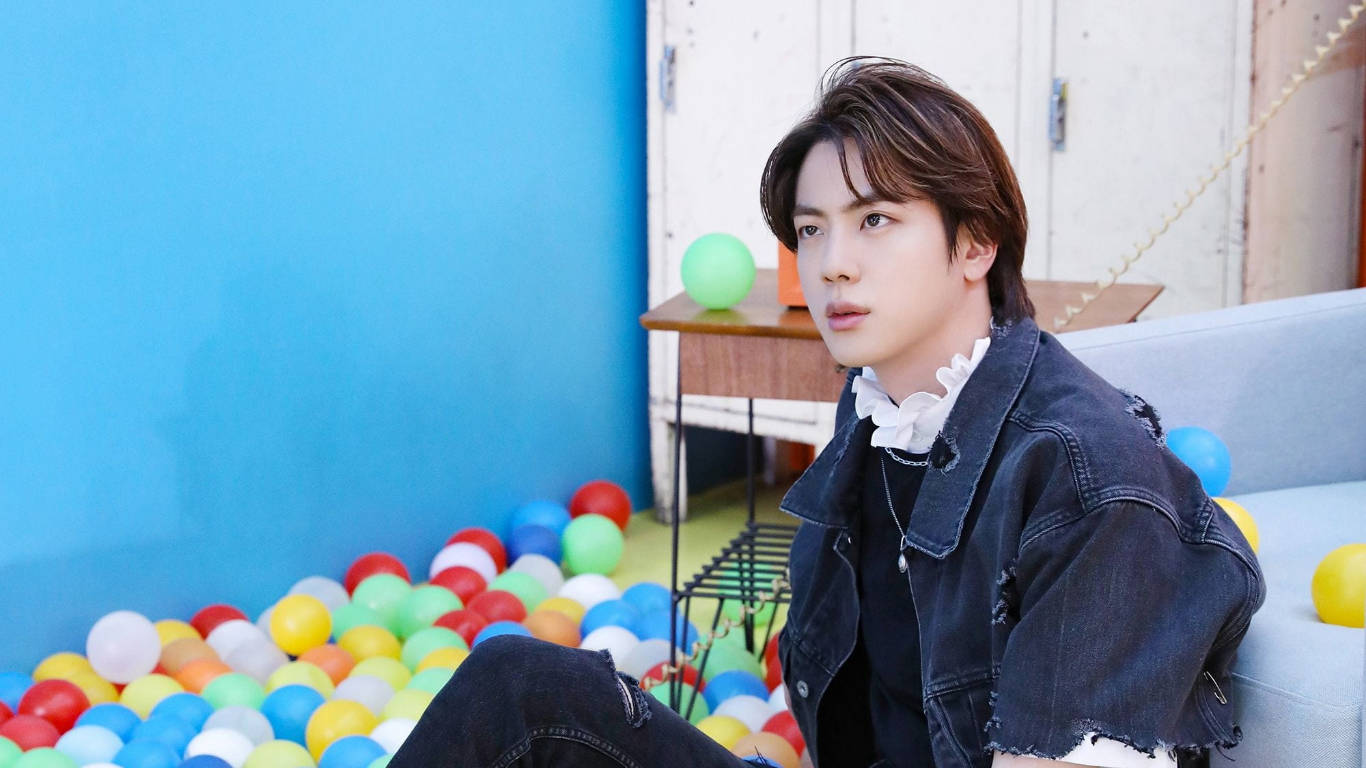 kim seok jin 桌布 壁紙 — 一位身穿牛仔外套的年輕男子坐在色彩繽紛的球池旁邊，背景是明亮的藍色牆壁。