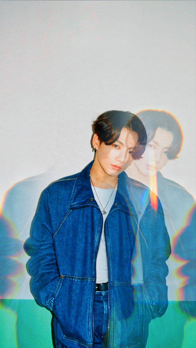 Jungkook 桌布 壁紙 — 一位身穿牛仔外套的年輕男子站在素色牆前，身後有色彩繽紛的彩虹鏡頭光暈效果。