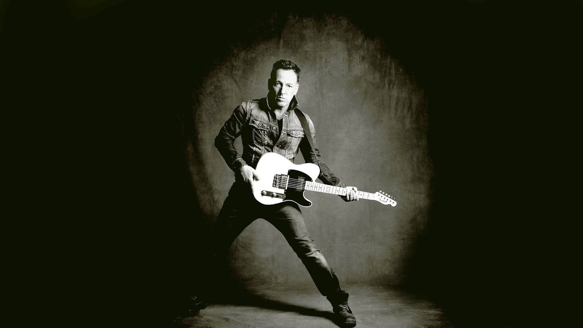 Bruce Springsteen 桌布 壁紙 — 一位身穿牛仔外套的音樂家在聚光燈下，以深色紋理背景為襯，彈奏白色電吉他。