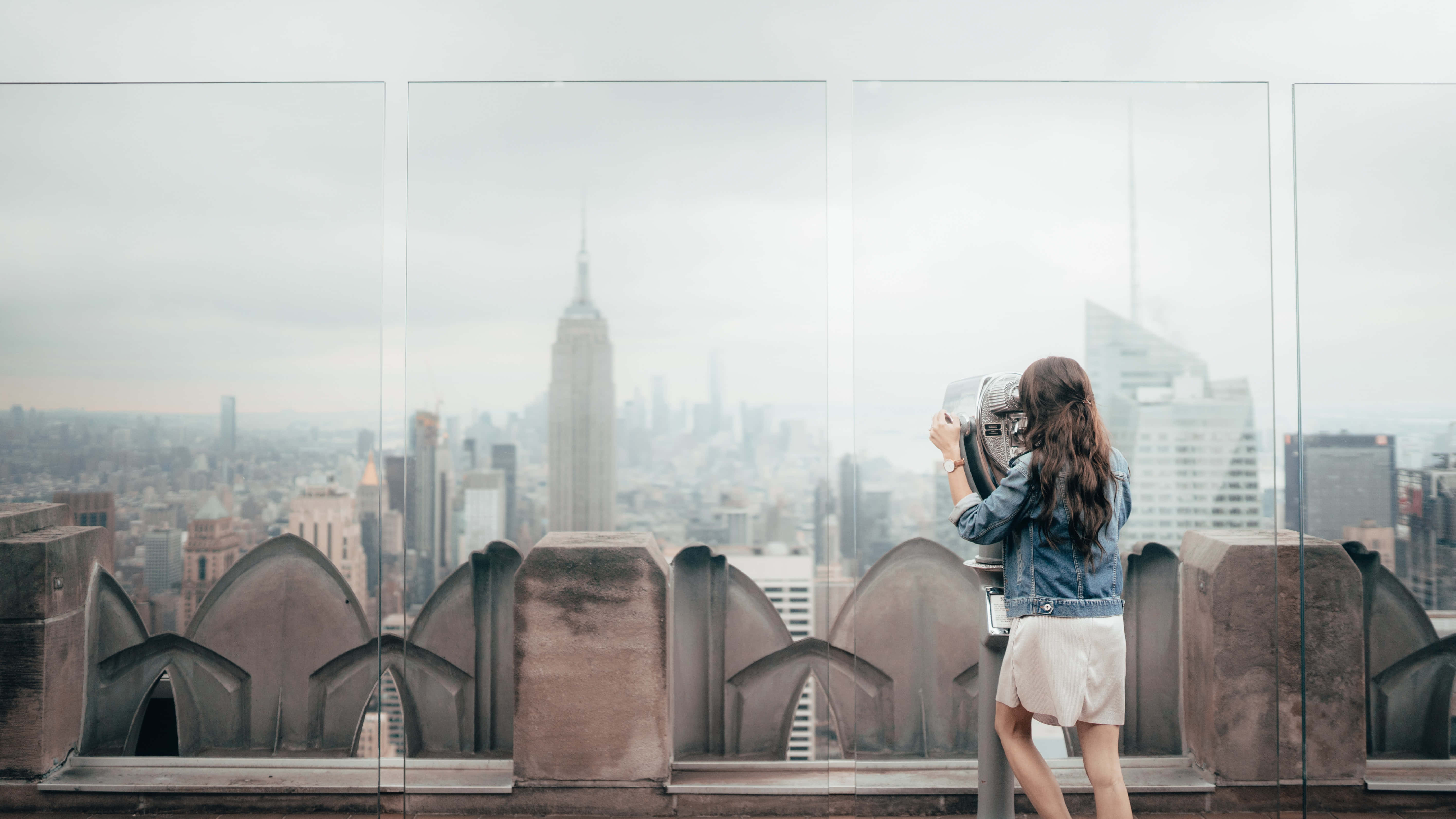 Nyc 桌布 壁紙 — 一位身穿牛仔夾克的女性在設有拱形石欄杆的屋頂上，使用投幣式望遠鏡眺望紐約市的天際線。