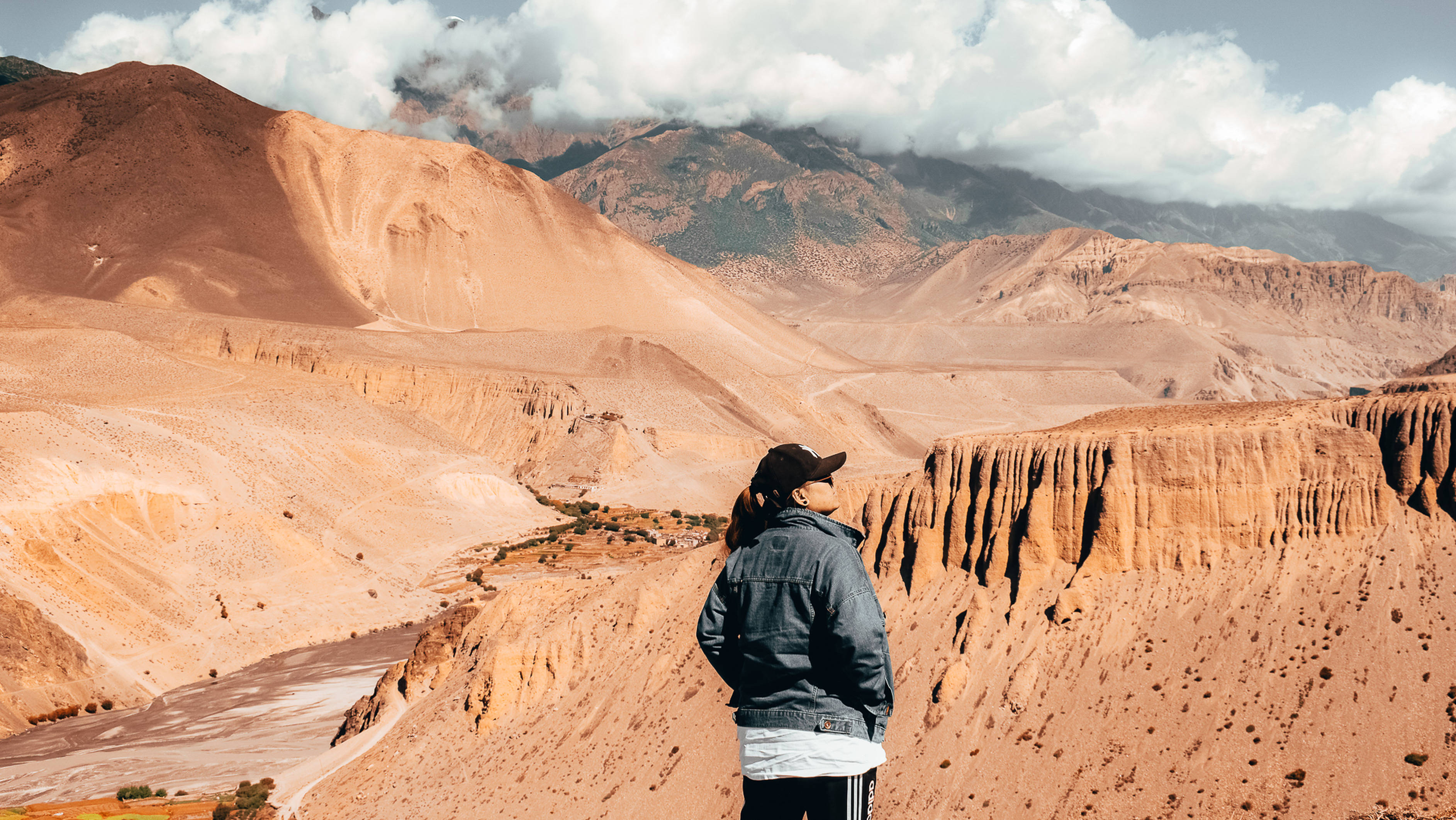 Nepal 桌布 壁紙 — 一位身穿牛仔夾克的孤獨旅人站在崎嶇的峽谷邊緣，俯瞰著多雲天空下廣闊的橙紅色沙漠山谷。