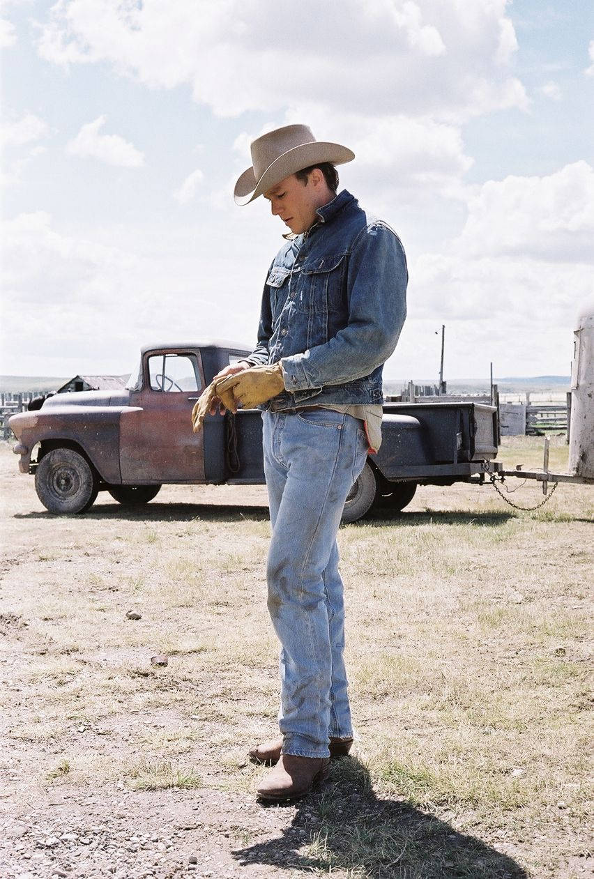 brokeback mountain 桌布 壁紙 — 一位身穿牛仔布和帽子的牛仔站在一輛生鏽的皮卡車旁，在明亮的陰天天空下，乾草上。