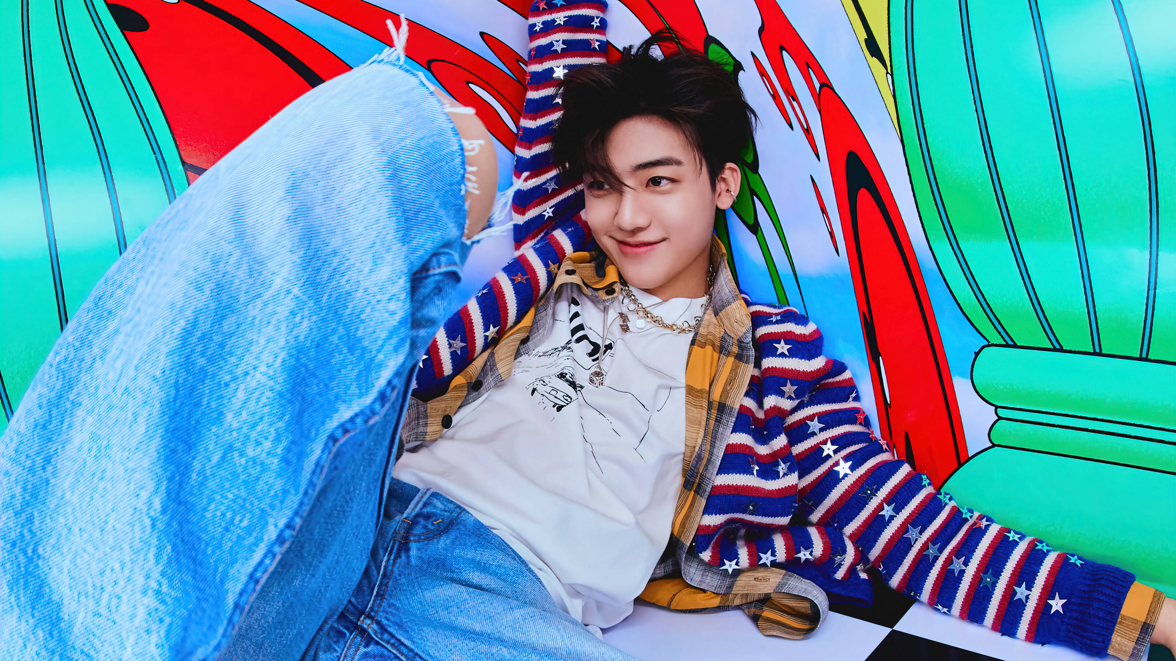 jaemin 桌布 壁紙 — 一位身穿牛仔布和星星圖案毛衣的年輕男子靠在一幅色彩鮮豔、風格卡通、帶有強烈紅、綠、藍色的壁畫上。