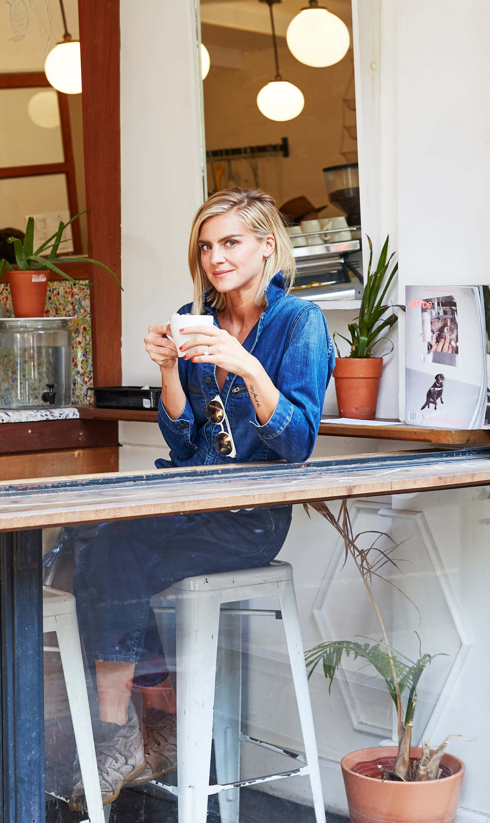 eliza coupe 桌布 壁紙 — 一位身穿牛仔布的女性坐在咖啡館櫃檯的凳子上，手持咖啡杯，周圍有溫暖的燈光和盆栽植物。