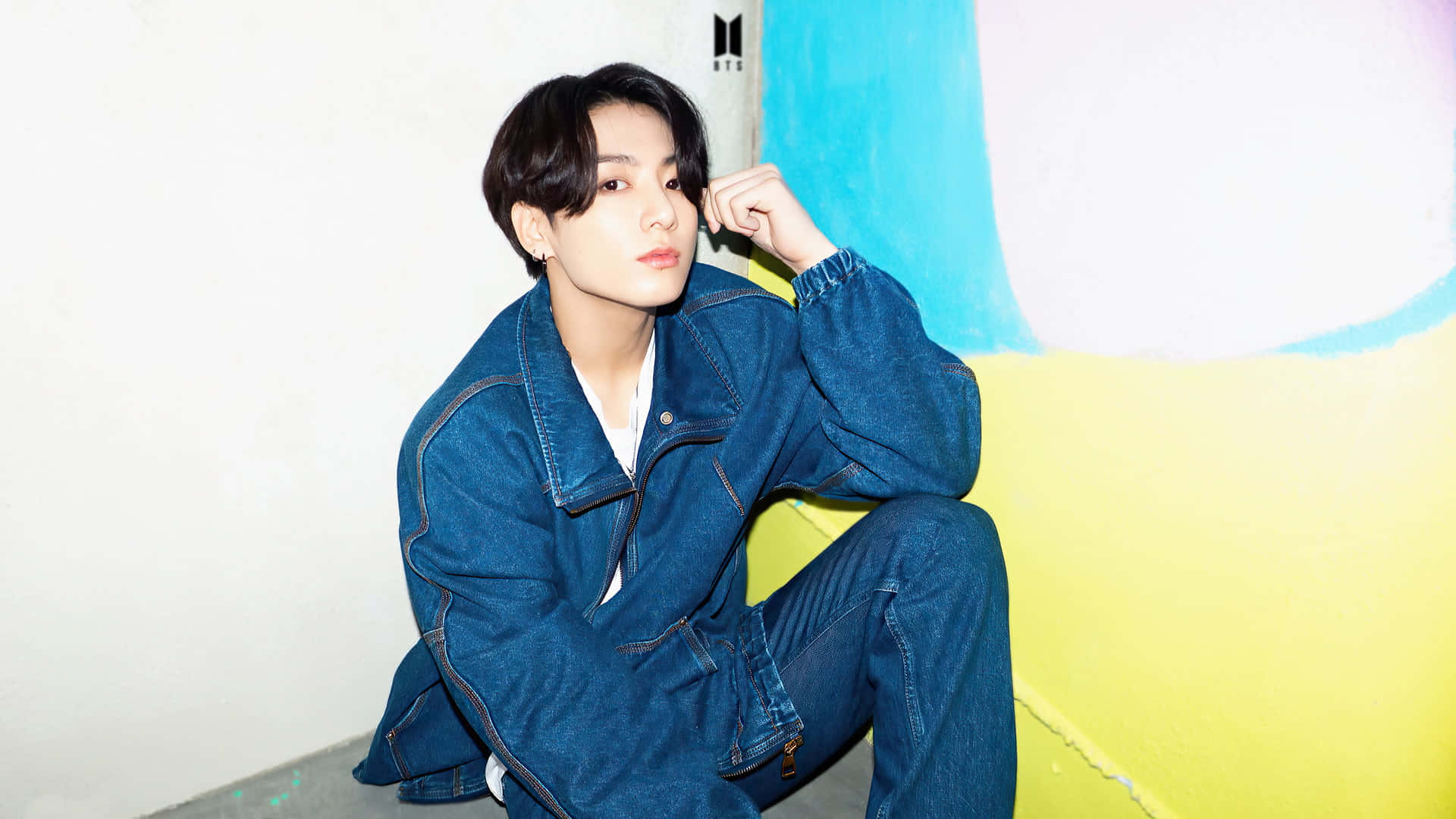Jungkook 桌布 壁紙 — 一位身穿牛仔裝的年輕男子隨意地坐在色彩繽紛的牆邊，牆上有大膽的黃色和藍色區域。