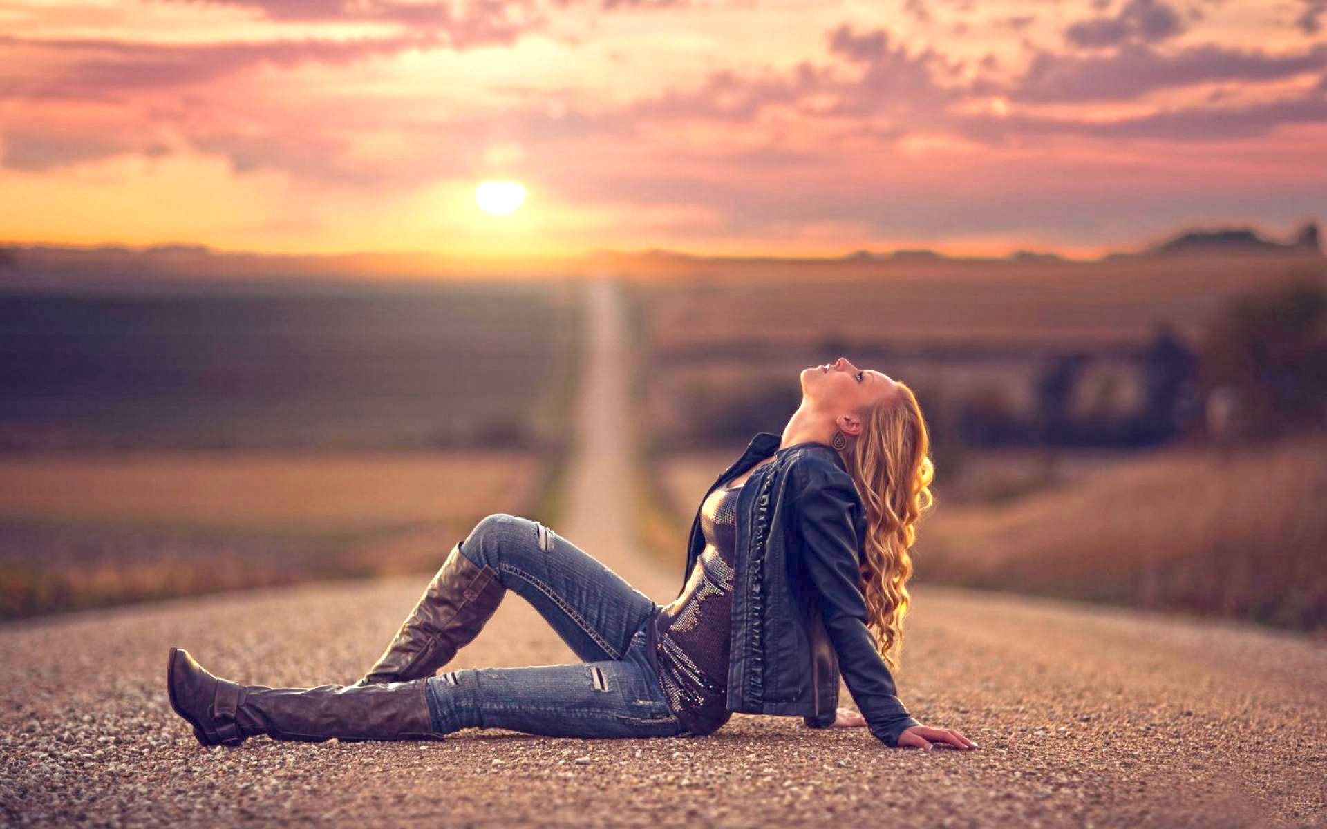 Sad Girl 桌布 壁紙 — 一位身穿牛仔褲和靴子的女士坐在日落時分的鄉村道路上，頭向後仰，閉上眼睛，沐浴在溫暖的金色光芒中。
